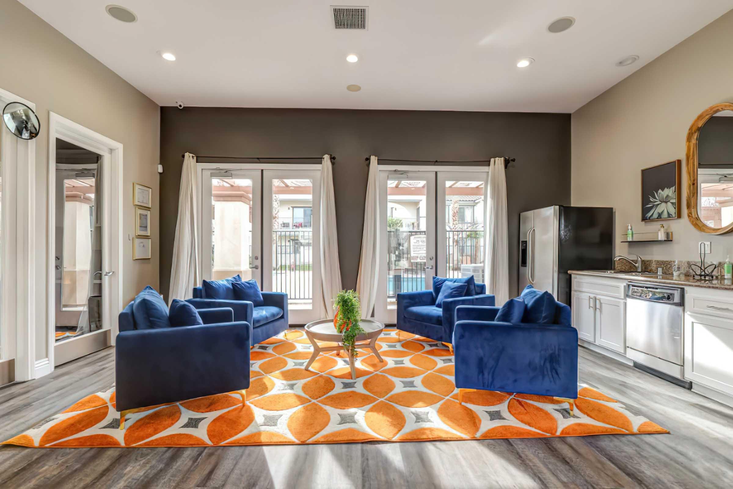 A modern lounge area featuring four dark blue upholstered chairs arranged around a circular coffee table on an orange and white patterned rug. Large windows with sliding doors let in natural light, and there's a kitchenette visible in the background. The walls are painted in neutral tones, creating a cozy atmosphere.
