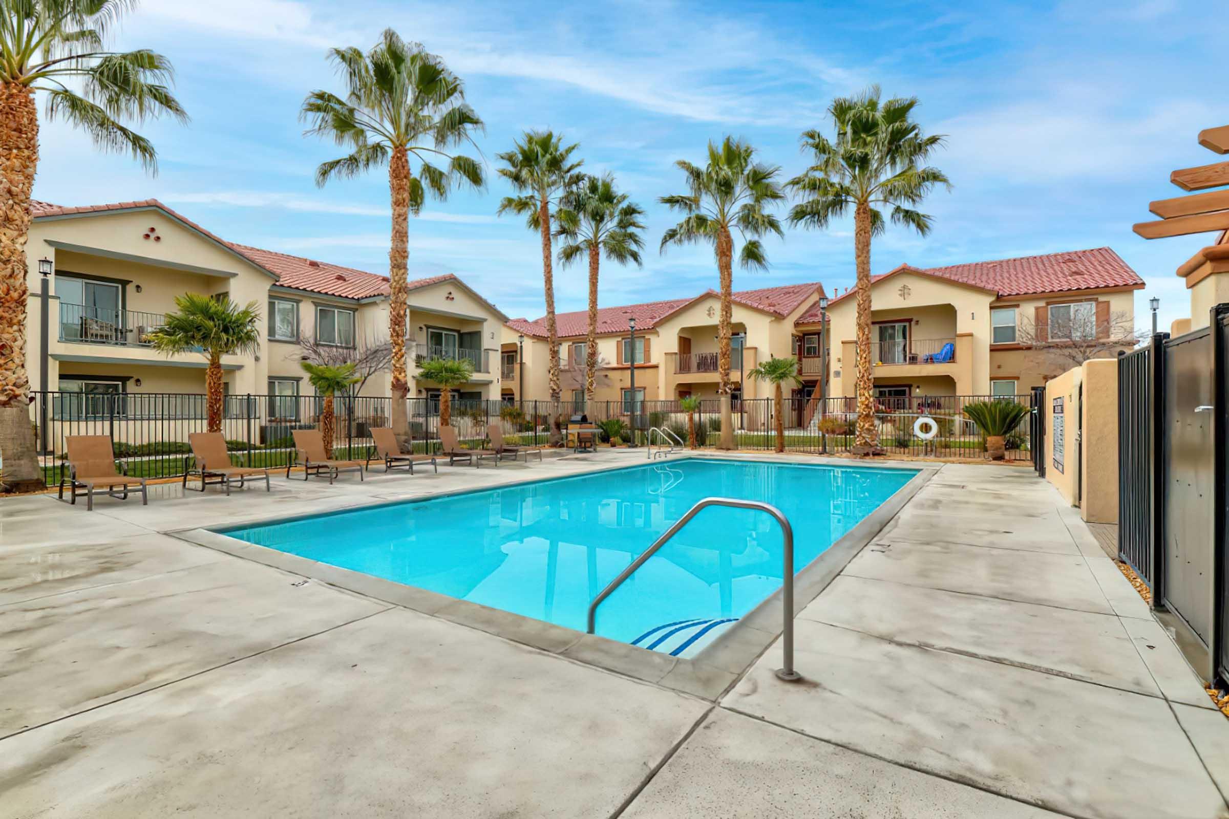 A serene outdoor pool area surrounded by palm trees and lounge chairs, situated in front of multi-story apartment buildings. The pool is bright blue and inviting, set against a clear sky and landscaped greenery. The scene conveys a relaxing, resort-like atmosphere.
