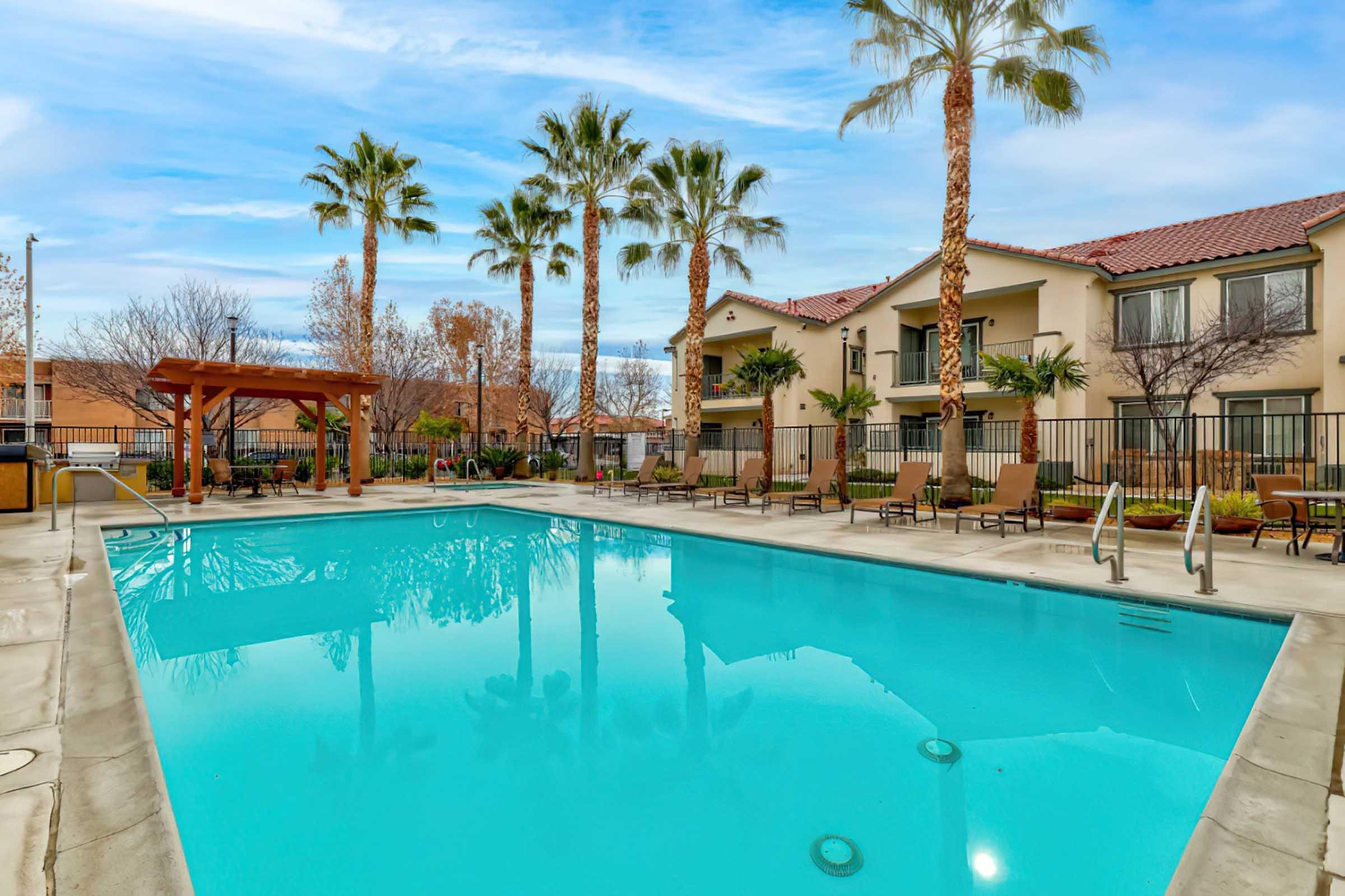 A clear blue swimming pool surrounded by palm trees and lounge chairs. In the background, there are two-story residential buildings with balconies. The sky is partly cloudy, adding to the serene atmosphere.