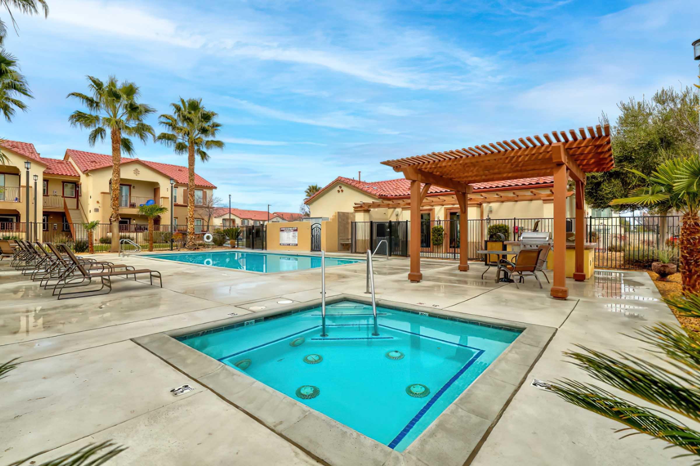 A sunny outdoor pool area featuring a large swimming pool and a smaller jacuzzi. Surrounding the pools are lounge chairs and palm trees, with a pergola providing shade. In the background, there are residential buildings, creating a relaxing atmosphere. The sky is clear with a few clouds.
