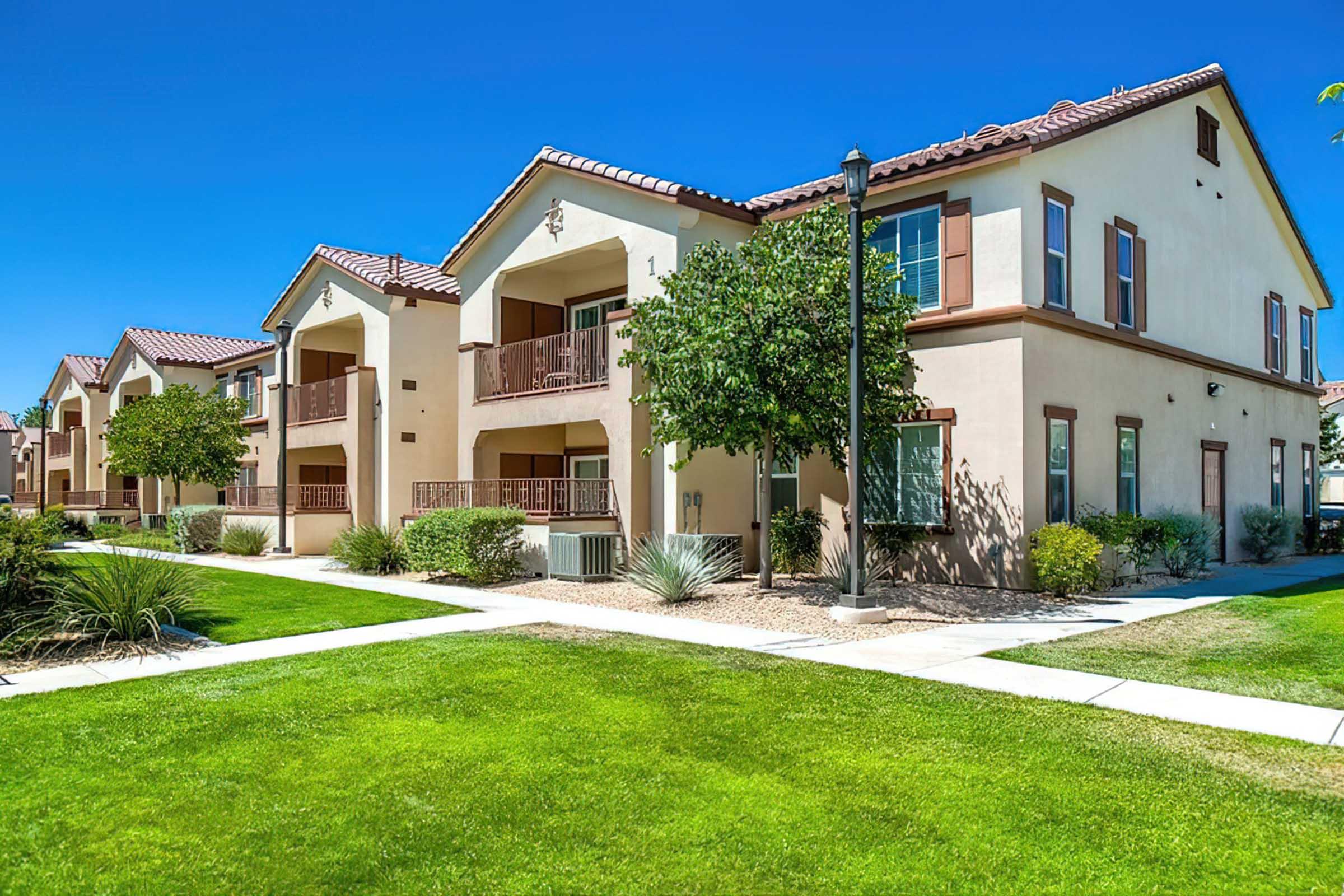 A row of modern, two-story apartment buildings with beige and brown exteriors. The buildings feature balconies, surrounded by well-maintained lawns, shrubs, and small trees. Clear blue skies and bright sunlight enhance the inviting atmosphere of the residential area.
