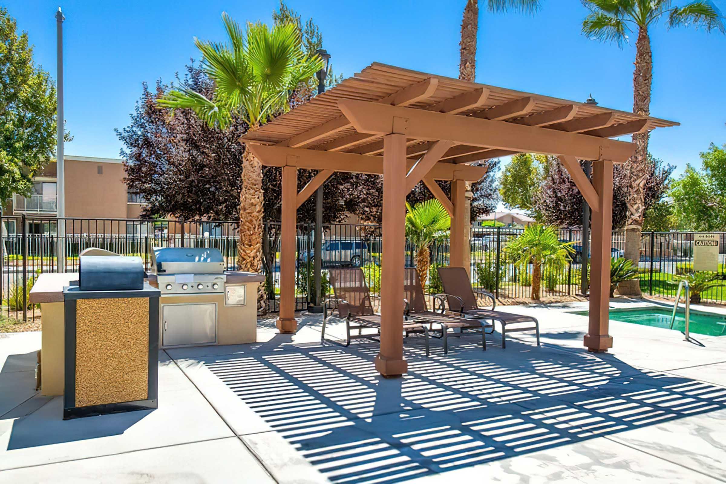 A shaded outdoor area featuring a wooden pergola, a barbecue grill, and two lounge chairs. Palm trees and dark foliage surround the space, with a clear blue sky above. The concrete floor casts shadows in the sunlight, creating a relaxing atmosphere ideal for social gatherings or relaxation.
