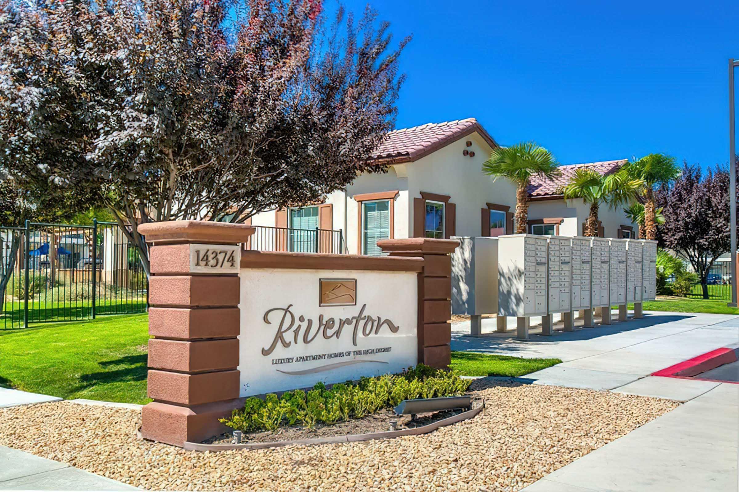 Signage for "Riverton," an apartment community, featuring the name prominently displayed. The building has a clean, modern design with landscaping that includes palm trees and decorative stones. There are mailbox units visible beside the entrance, all set against a clear blue sky.