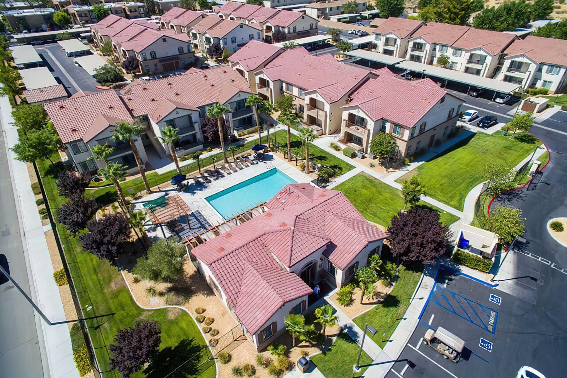 Aerial view of a residential complex featuring multiple tan buildings with red tile roofs. In the center, there is a shaped swimming pool surrounded by lounge chairs and palm trees, along with landscaped green areas and parking spaces visible. The setting appears sunny and well-maintained.