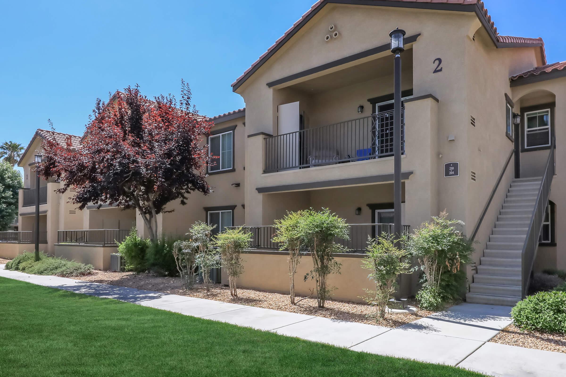 A well-maintained apartment complex featuring two-story buildings with balconies. Lush green grass and young shrubs line a paved walkway leading to the entrance. A decorative tree adds aesthetic charm to the landscape, and clear blue skies create a bright and welcoming atmosphere.