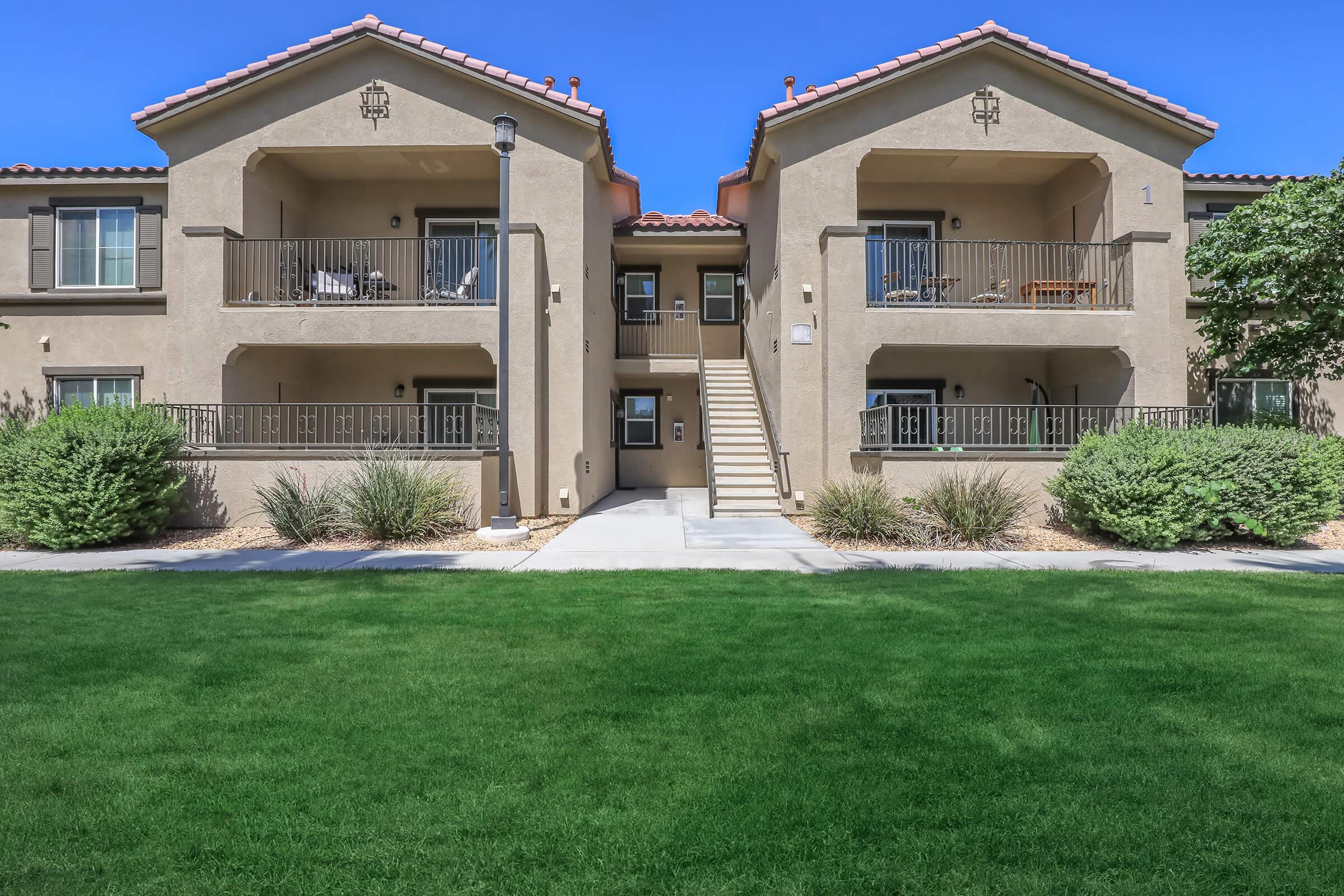 Two-story apartment building featuring a light beige exterior with balconies on each unit, surrounded by green grass and landscaping. A central pathway leads to the entrance stairs, with shrubs flanking the sides. The sky is clear and blue, providing a bright atmosphere.