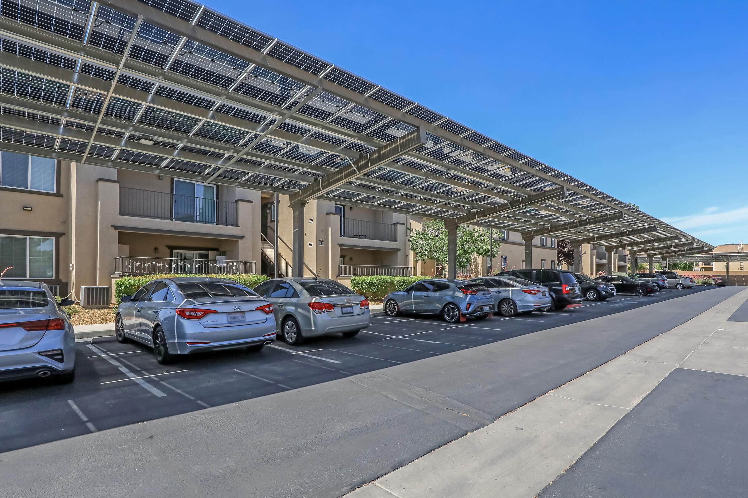 A row of parked cars under solar panel canopies next to a residential building. The clear blue sky provides a bright backdrop to the scene. The area is well-maintained, showcasing environmentally friendly energy solutions with multiple vehicles parked in designated spaces.