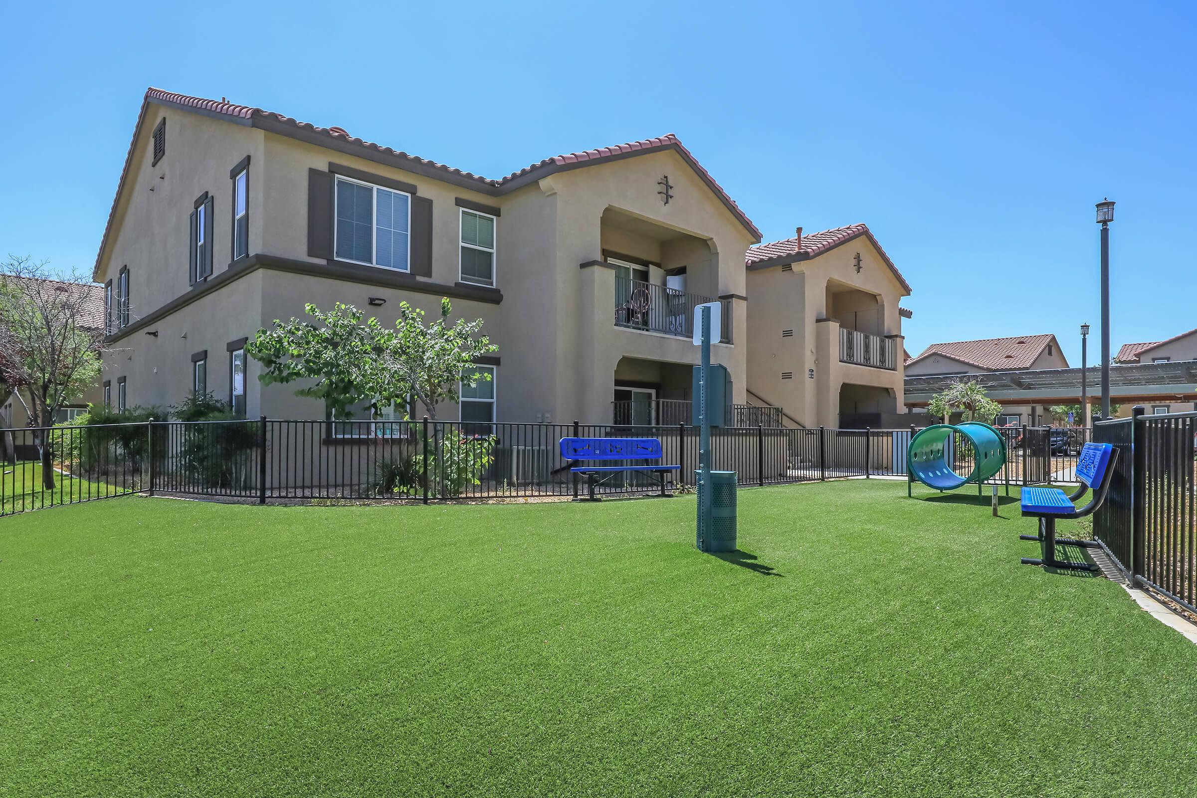 A well-maintained green lawn area in front of a two-story residential building. The space features a playground with swings and slides, surrounded by a black fence. Nearby, there are benches and trees, with bright blue skies above, indicating a sunny day.