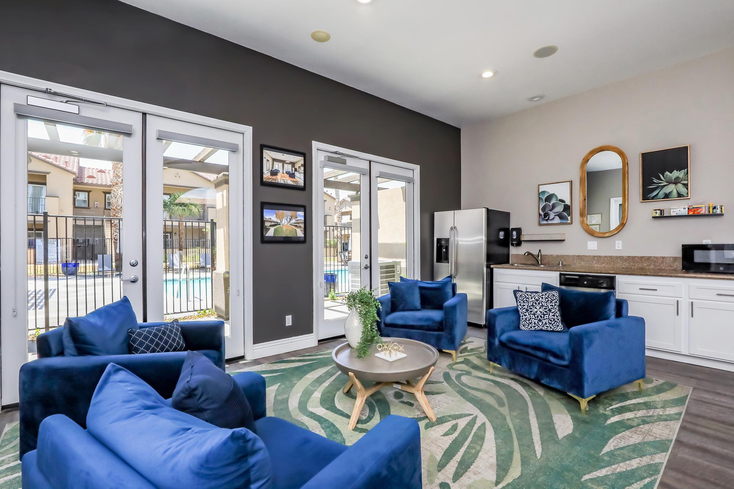 A modern living space featuring two blue armchairs and a round coffee table on a patterned rug. Large glass doors provide a view of a pool area outside. A kitchen with stainless steel appliances is visible in the background, along with decorative wall art and plants. The walls are painted in a dark and light color scheme.