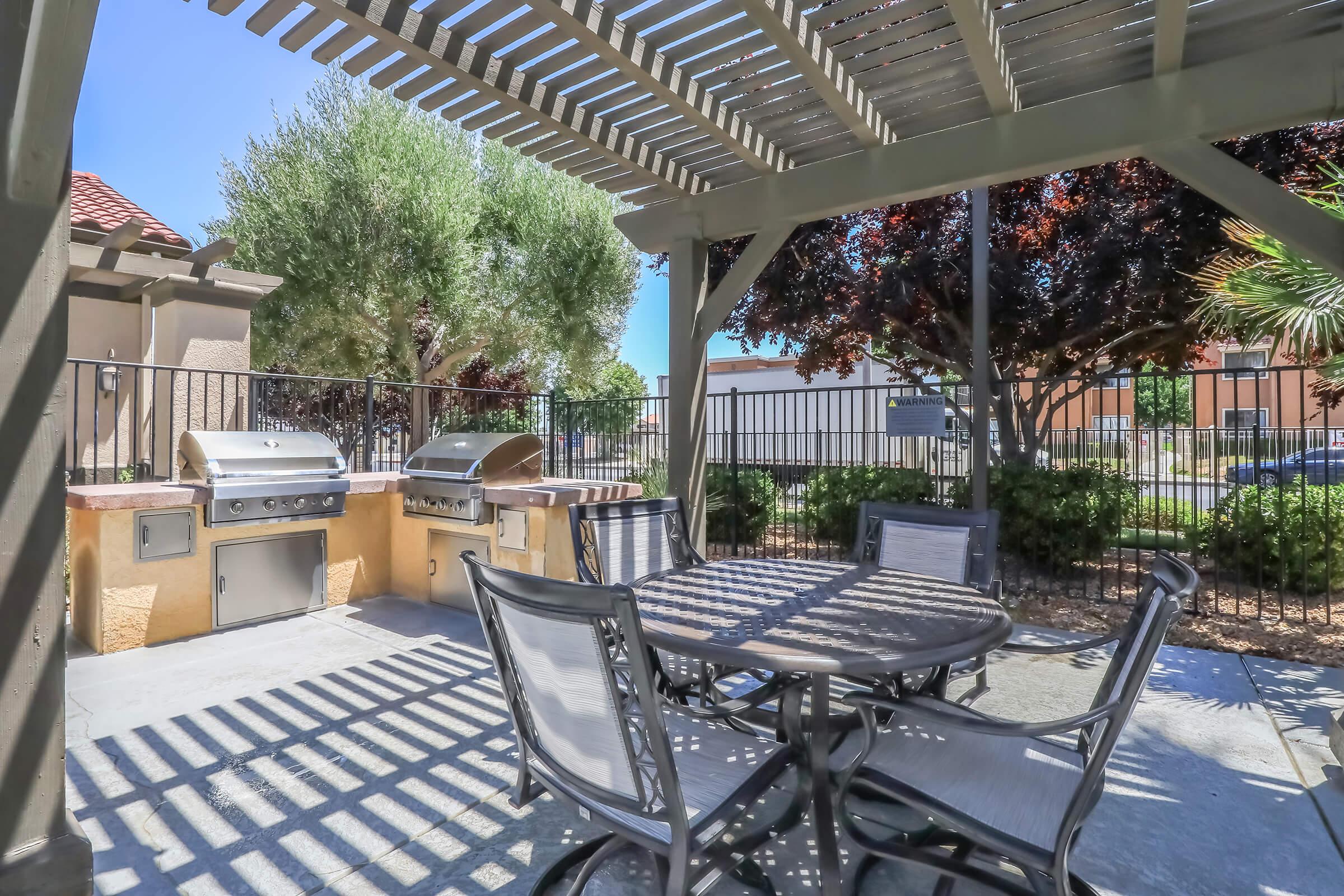 Outdoor dining area featuring a shaded pergola, a round table with chairs, and two built-in grills. Surrounding greenery adds a cozy atmosphere, with a fence in the background providing privacy. The scene is well-lit, suggesting a pleasant environment for gatherings or barbecues.