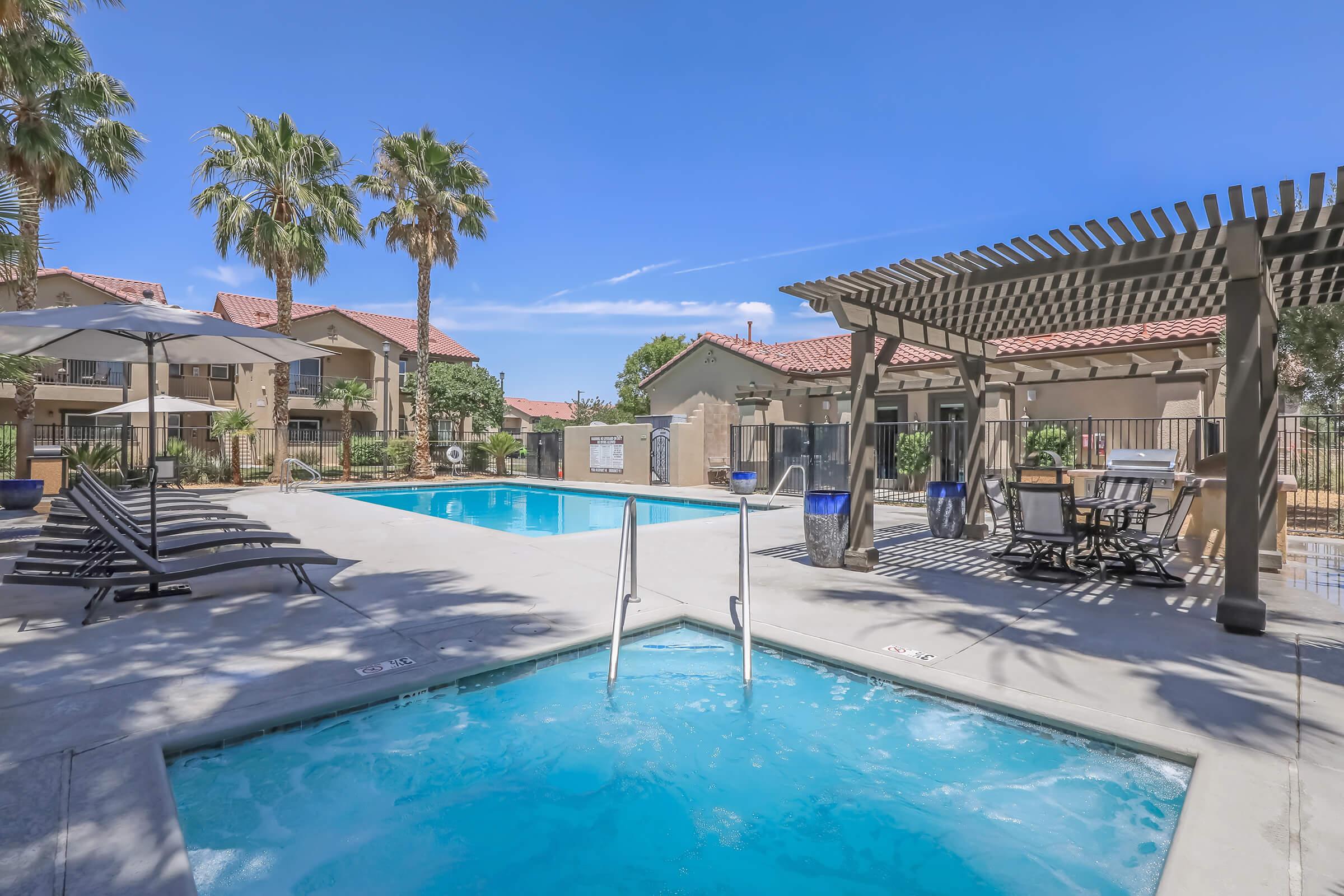 A resort-style swimming pool area featuring a larger pool and a hot tub, surrounded by lounge chairs and umbrellas. Palm trees provide shade, and a covered seating area with tables and chairs is visible. The scene is bright and sunny, ideal for relaxation and leisure.