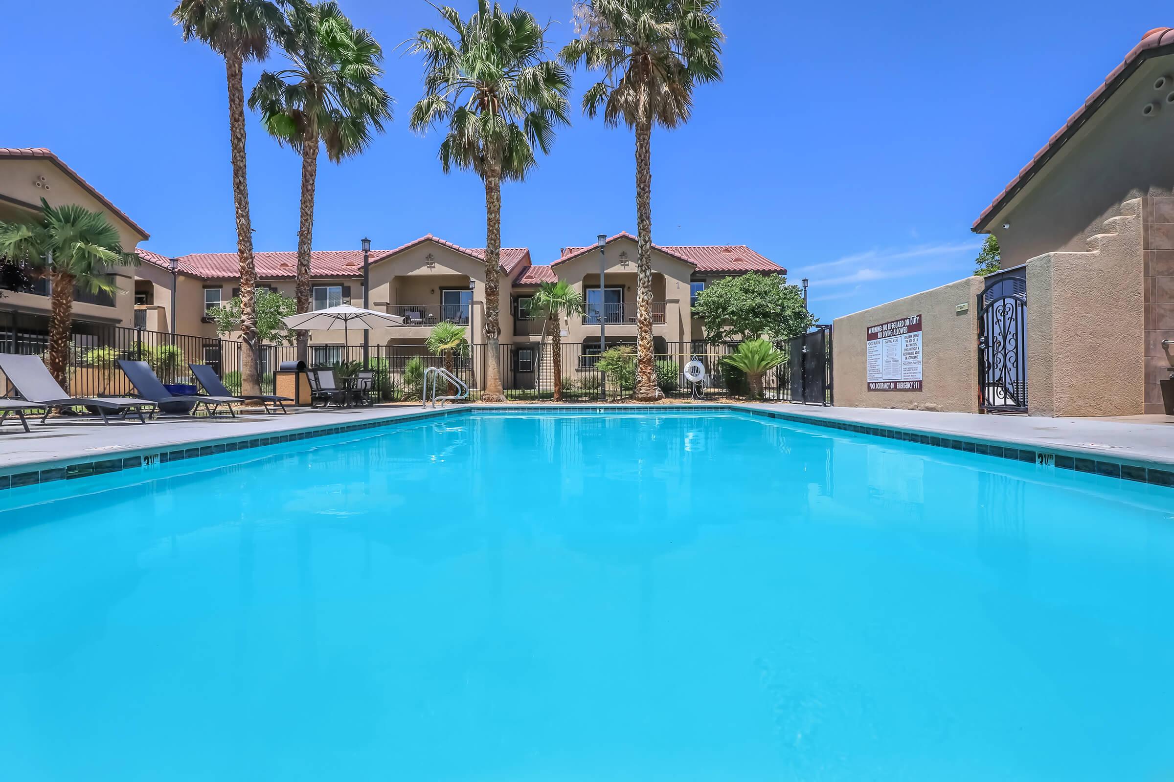 A clear blue swimming pool surrounded by palm trees and lounge chairs. In the background, there are residential buildings with a sunny sky overhead. The scene conveys a relaxed, inviting atmosphere typical of a resort or apartment complex.