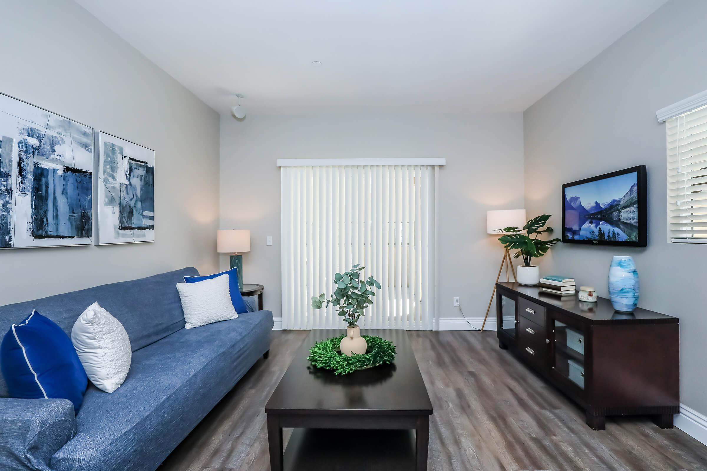 A modern living room featuring a gray sofa with blue and white pillows, a wooden coffee table with a decorative plant centerpiece, and a media console. Light gray walls complement the space, which is illuminated by floor lamps. Large vertical blinds partially cover the window, and an art piece is displayed on the wall.