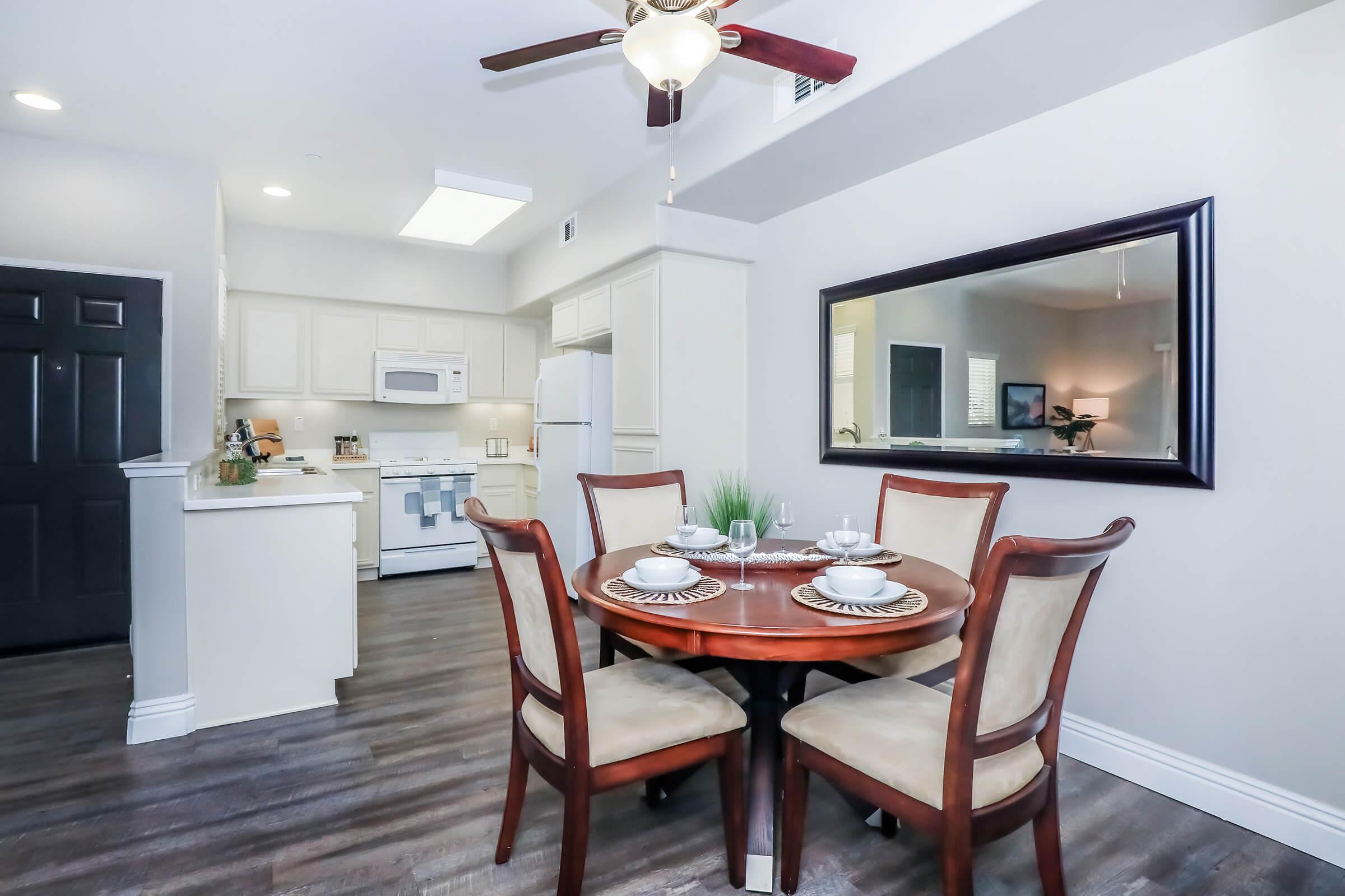 A bright and modern kitchen with white cabinetry and appliances. A small dining table set for four with elegant plates and silverware. The space features a ceiling fan, and a large mirror hanging on the wall adds depth to the room. Wood-like flooring complements the contemporary design.