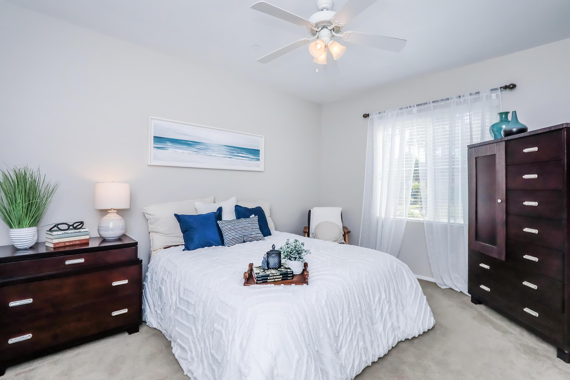 A cozy bedroom featuring a queen-sized bed with white bedding and blue accents, a nightstand with a lamp, a dresser with multiple drawers, and a large window with sheer curtains. Decorative elements include a vase with greenery and nautical-themed artwork on the wall.