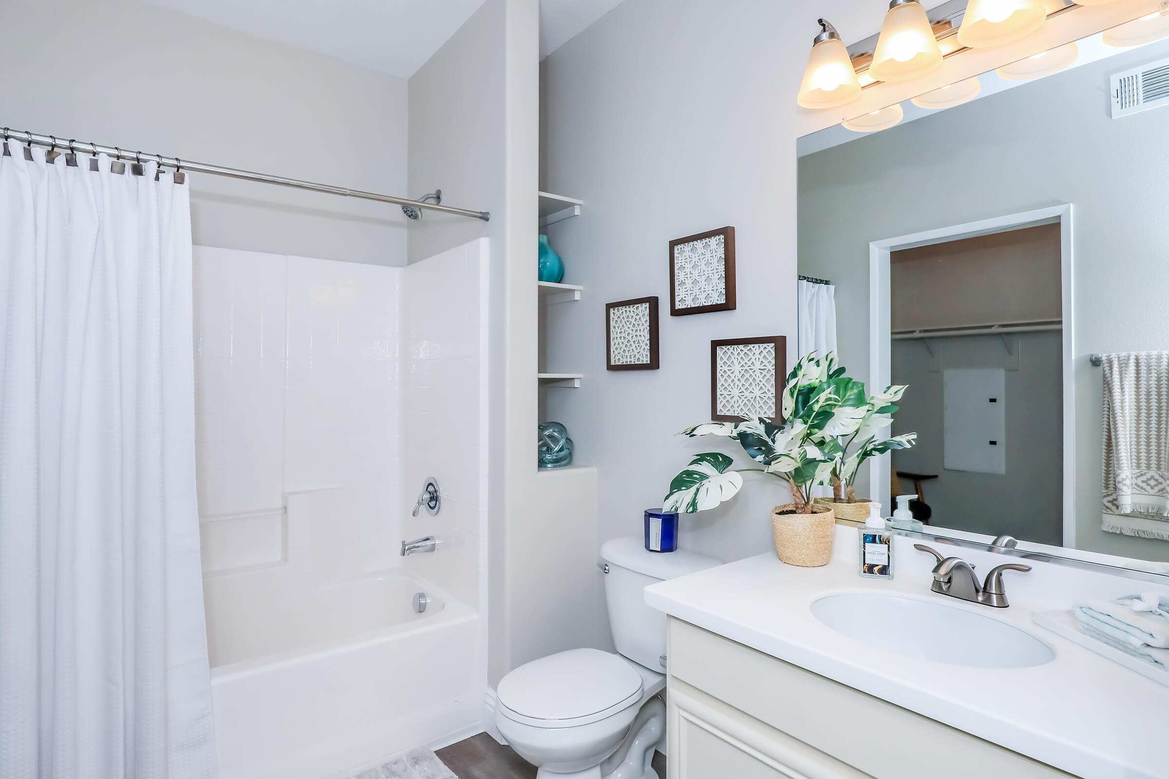 A clean and modern bathroom featuring a white bathtub with a shower curtain, a wall-mounted mirror with warm lighting, a sink with neatly arranged toiletries, and decorative plants. Shelves hold decorative items, while neutral-colored walls enhance the spacious feel of the room.