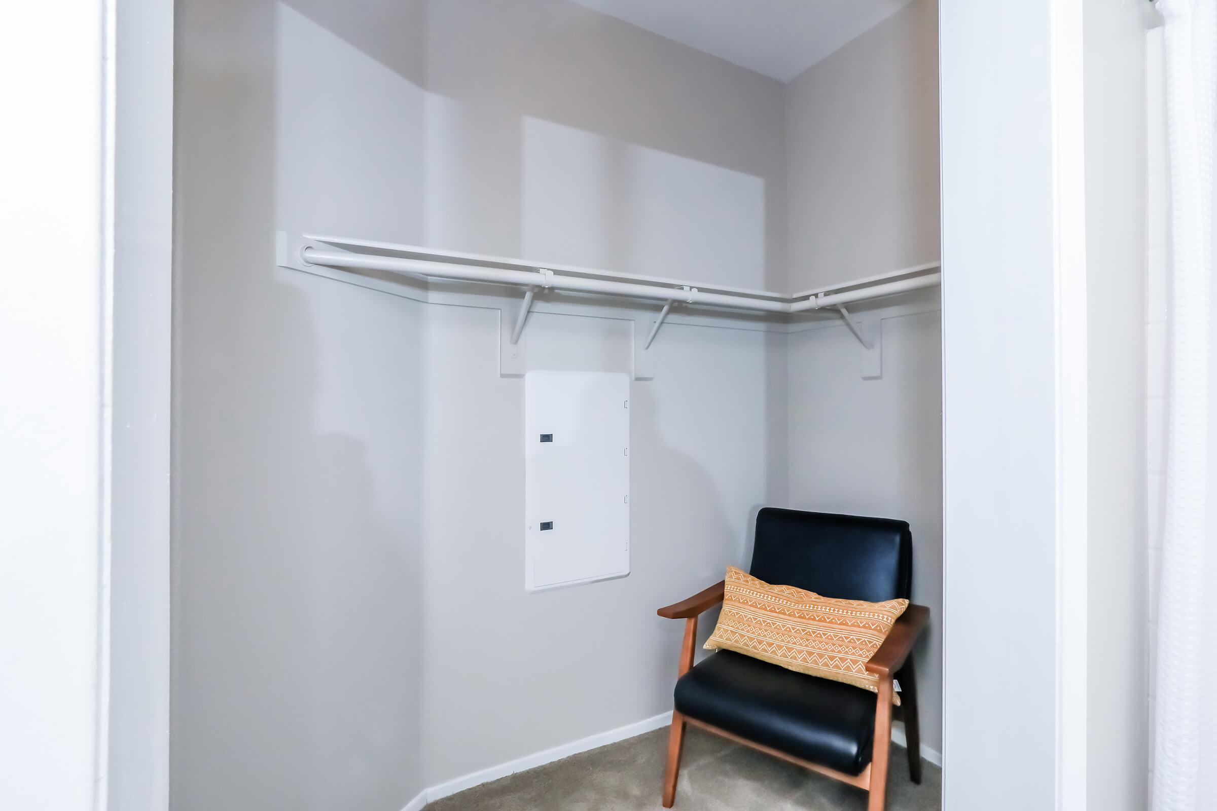 A minimalist closet with beige walls featuring two hanging shelves. There is a small chair with a black upholstered seat and wooden legs, adorned with an orange decorative pillow. A white electrical panel is mounted on the wall, and the carpeted floor is visible.