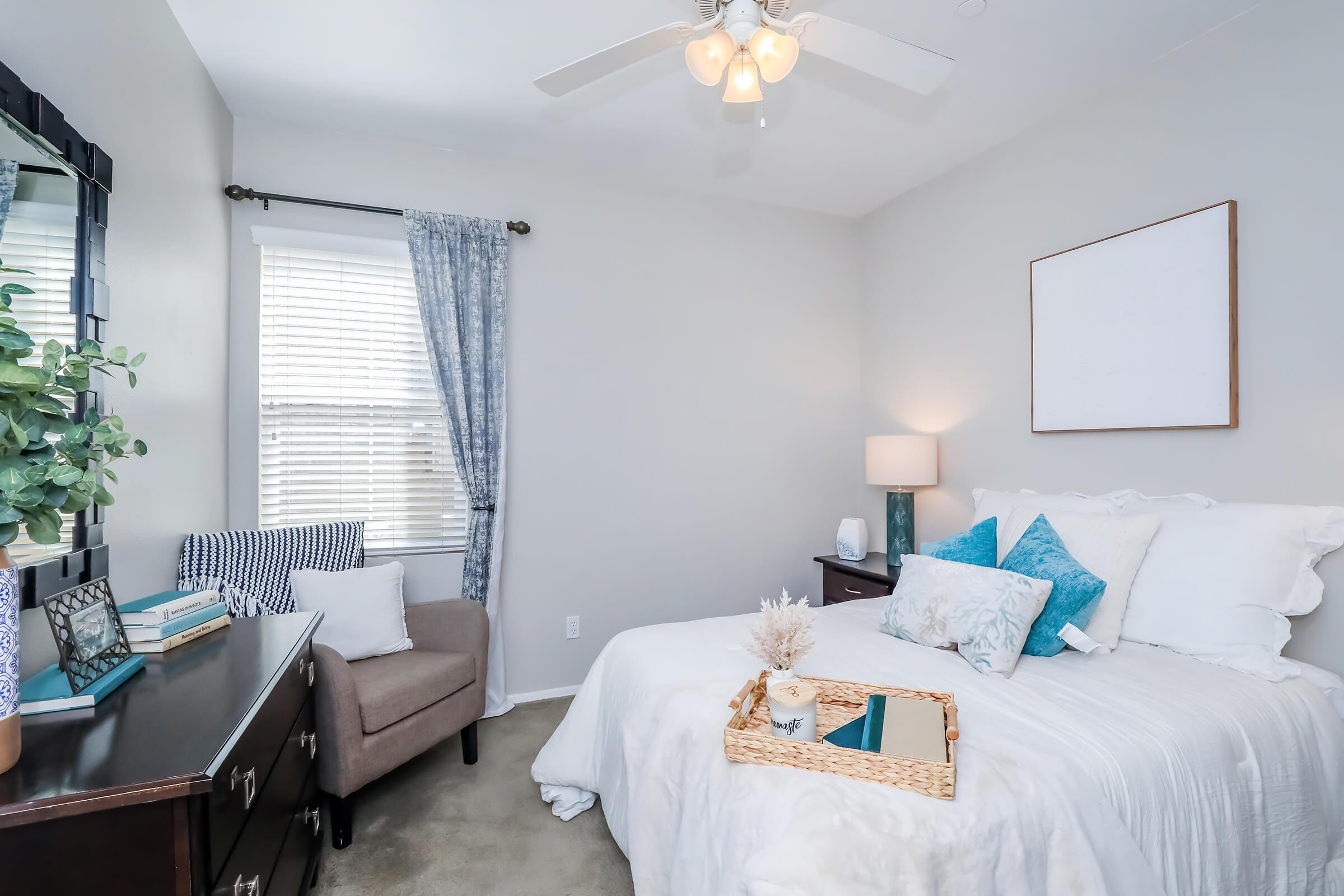 A cozy bedroom featuring a queen-sized bed with white linens and blue accent pillows. There's a stylish nightstand with a lamp, a chair in the corner, and a decorative plant on a dresser. Natural light streams in through a window with sheer curtains, creating a warm atmosphere.