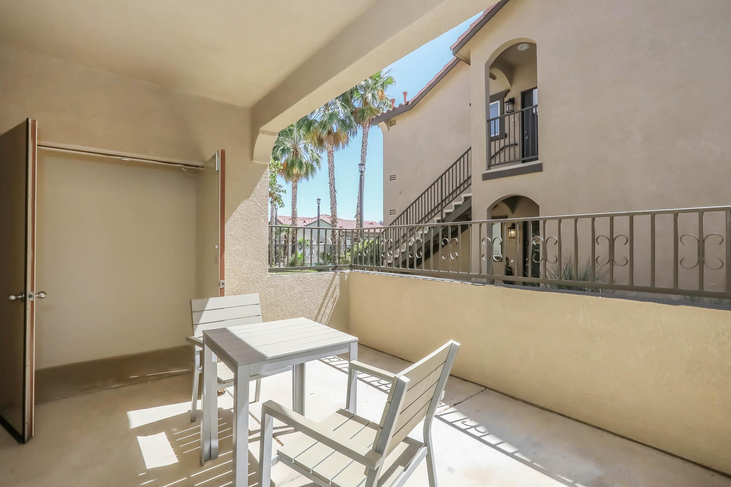 A patio area with a small table and chairs, surrounded by beige walls. There is an entrance to a room on the left, and in the background, a staircase leads to the upper level of the building. Palm trees are visible nearby, with bright blue sky overhead, indicating a warm and inviting outdoor space.