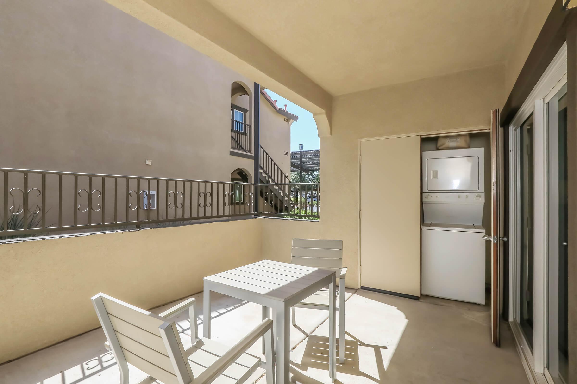 A small outdoor patio area features a table with four chairs, a laundry closet with washer and dryer, and a view of a neighboring building. The space is well-lit with beige walls and a railing, creating a cozy and functional area for relaxation or laundry access.