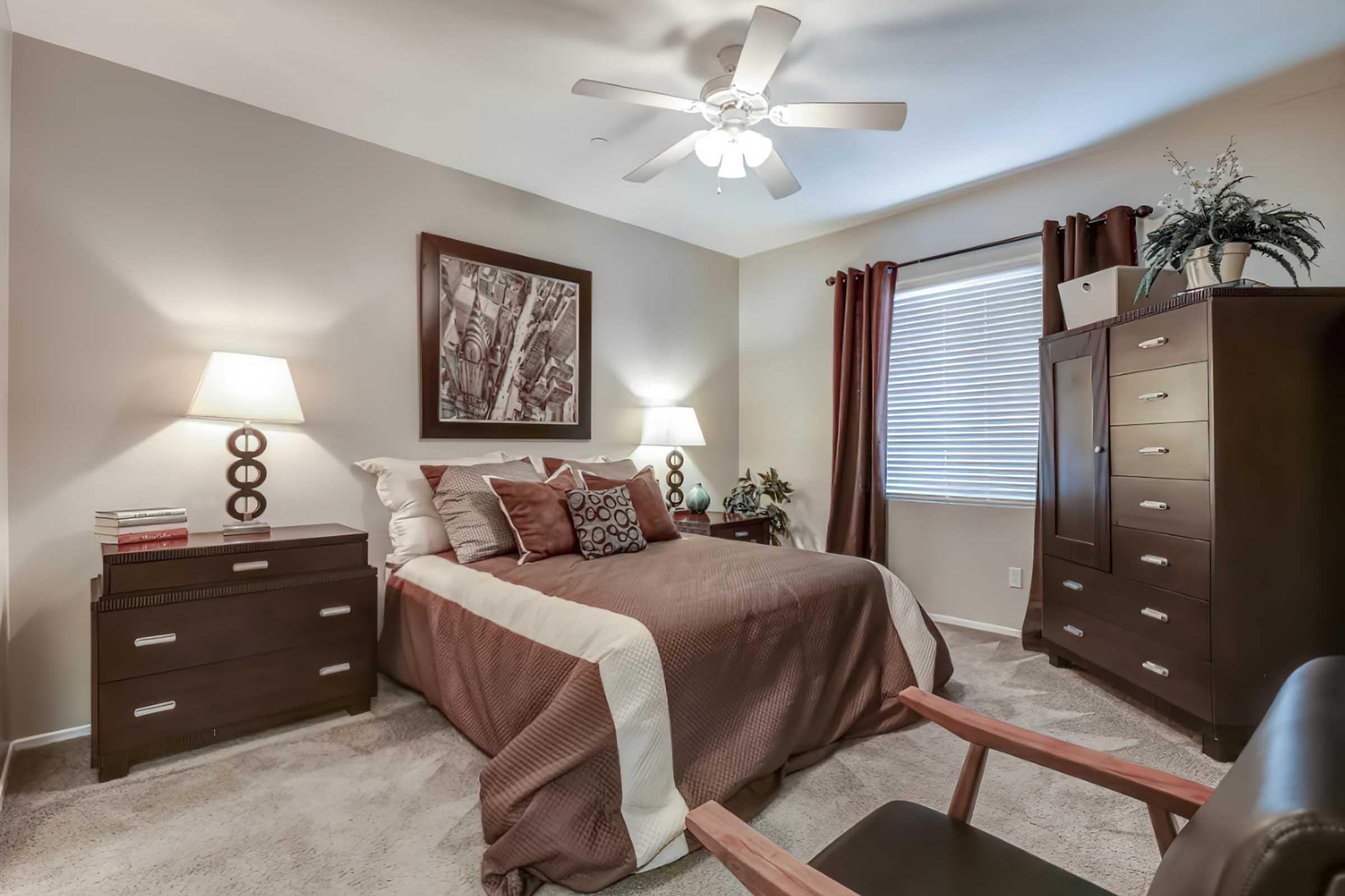 Cozy bedroom featuring a queen-sized bed with brown bedding, two bedside lamps, and a wooden dresser. A small armchair is in the corner, and framed artwork decorates the wall. Large window with blinds provides natural light, and potted plants add a touch of greenery. Neutral wall colors enhance the inviting atmosphere.
