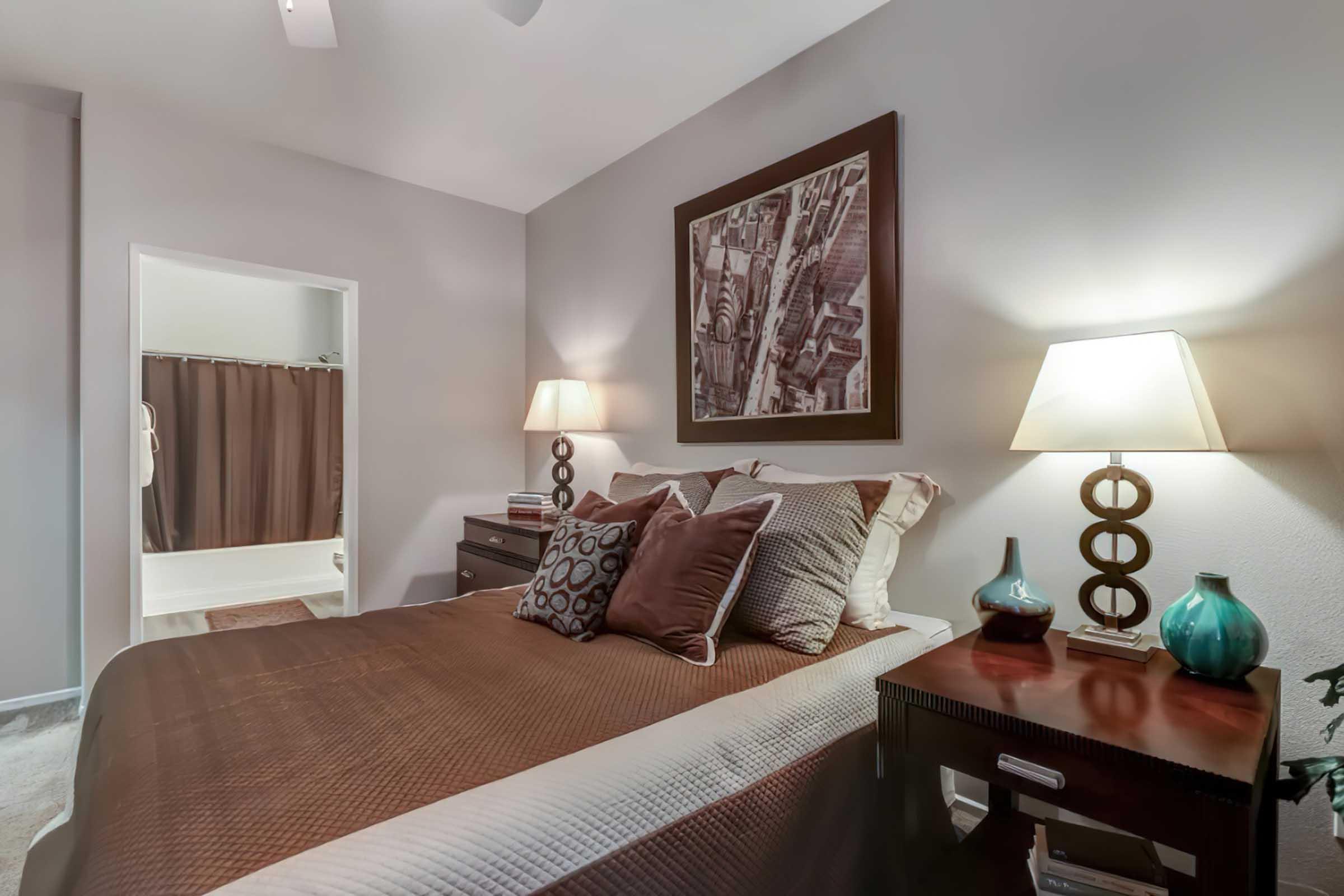 Cozy bedroom featuring a neatly made bed with decorative pillows, two bedside lamps, and a contemporary artwork above the headboard. A nightstand with a decorative vase sits next to the bed. A bathroom doorway is visible in the background, with a dark shower curtain. Soft, neutral tones create a relaxing atmosphere.