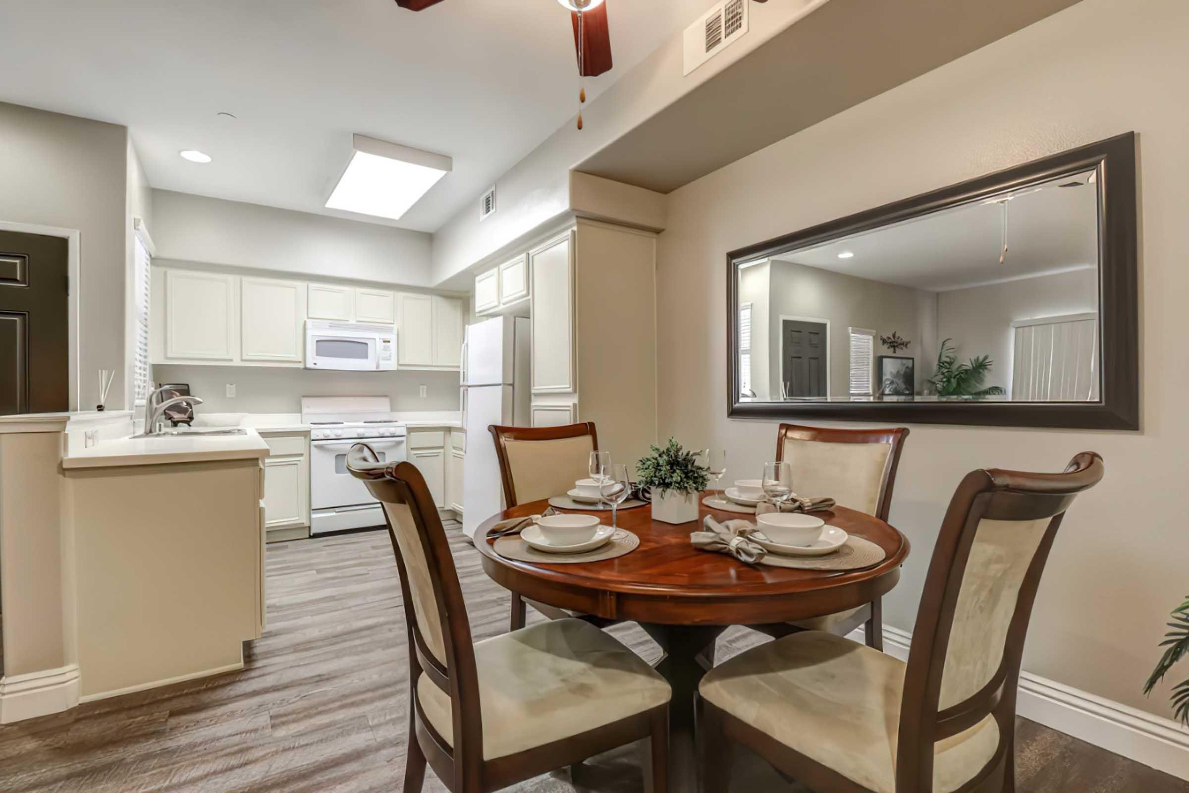 A cozy dining area featuring a round wooden table set with plates and utensils, surrounded by four upholstered chairs. In the background, a modern kitchen with white cabinets, a microwave, and a refrigerator, complemented by a large mirror on the wall, creating an inviting atmosphere.