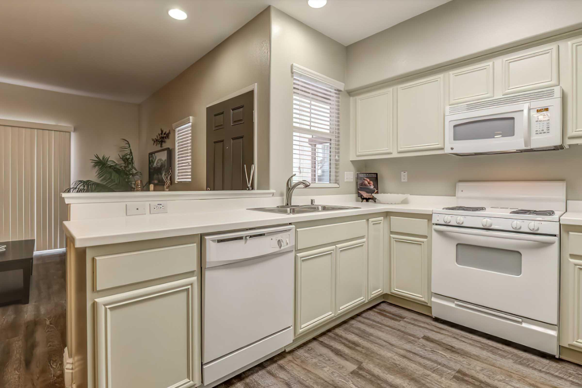 A modern kitchen featuring light-colored cabinets, a white countertop, a sink, a dishwasher, and a gas stove. Natural light enters through a window, and there's a sliding door partially visible. The floor is wooden, contributing to a warm, inviting atmosphere.