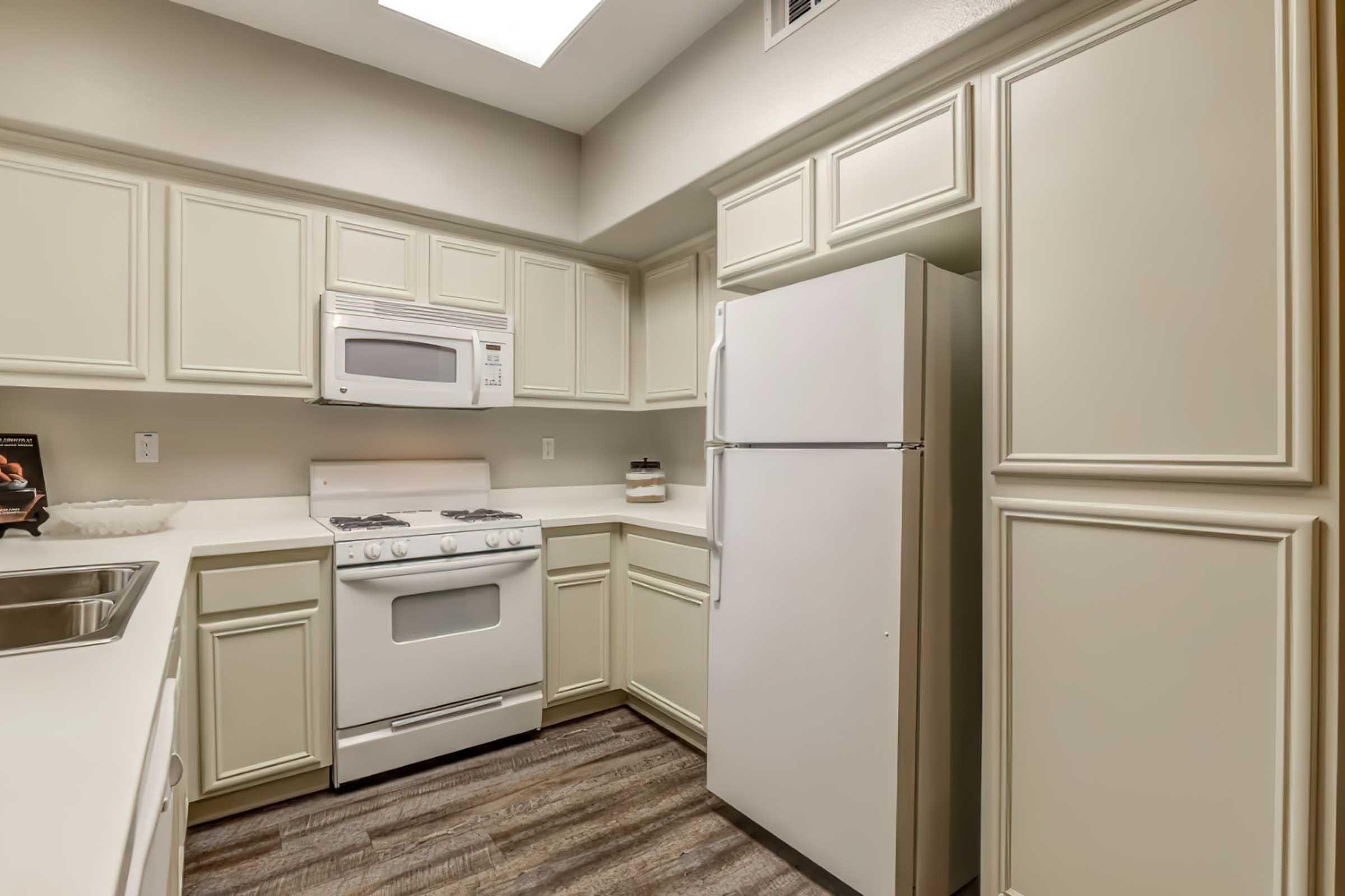 A modern kitchen featuring light-colored cabinets, a white gas stove, a microwave above the stove, and a white refrigerator. The countertops are light and clean, with a sink and minimal decorative items. The flooring is a wood-like laminate, creating a warm and inviting atmosphere.