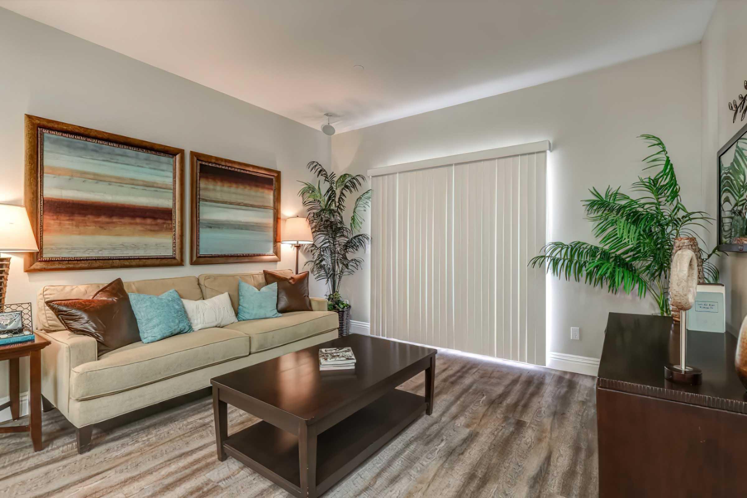 A cozy living room featuring a beige sofa with decorative pillows, two abstract paintings above it, and a wooden coffee table. There are indoor plants and a vertical blind window, providing a soft, inviting atmosphere with warm lighting.
