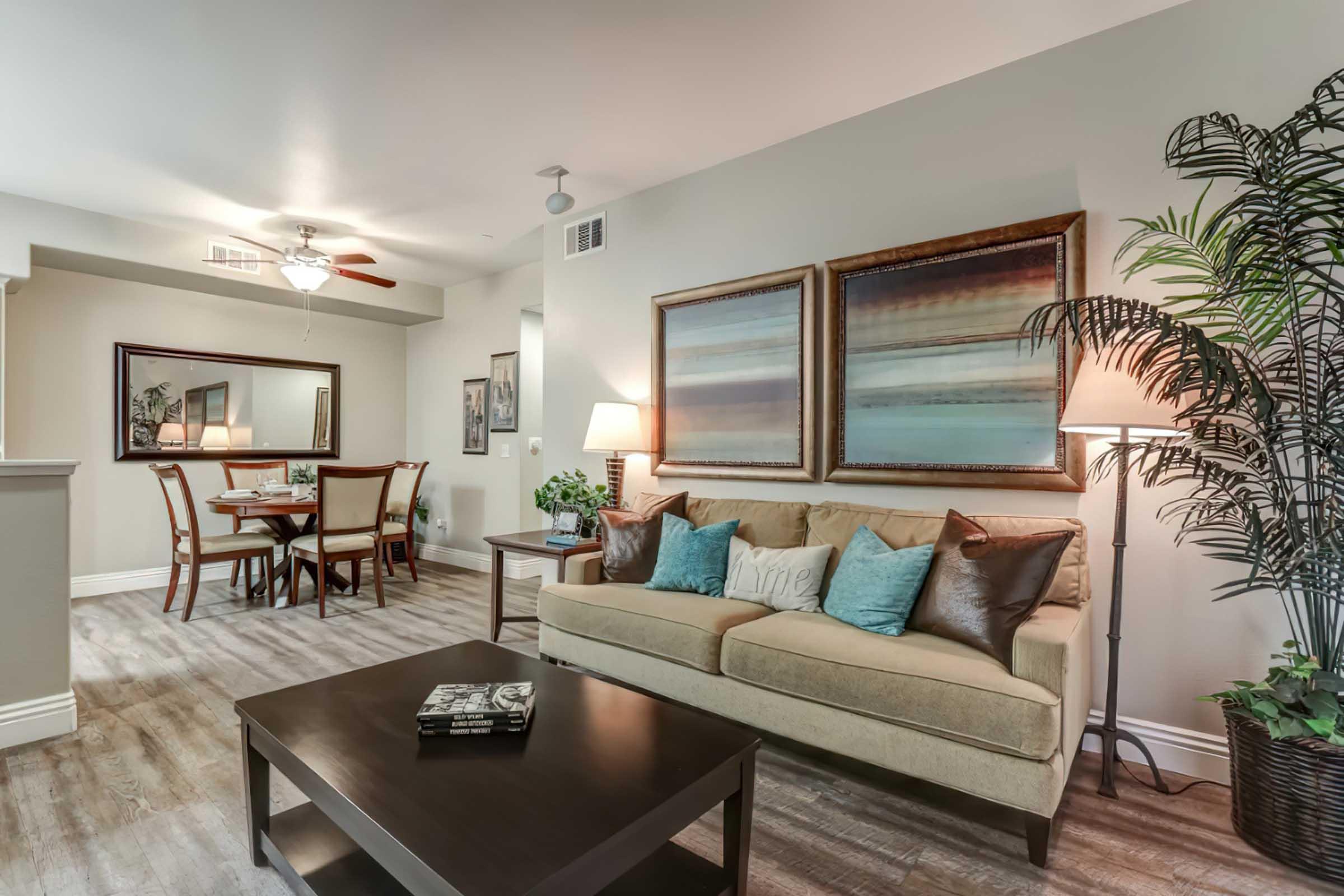 A cozy living room featuring a beige sofa adorned with turquoise and brown cushions. A dark wood coffee table sits in front. In the background, a dining area with a wooden table and chairs, illuminated by soft light. The walls are decorated with abstract art, and a potted plant adds a touch of greenery.