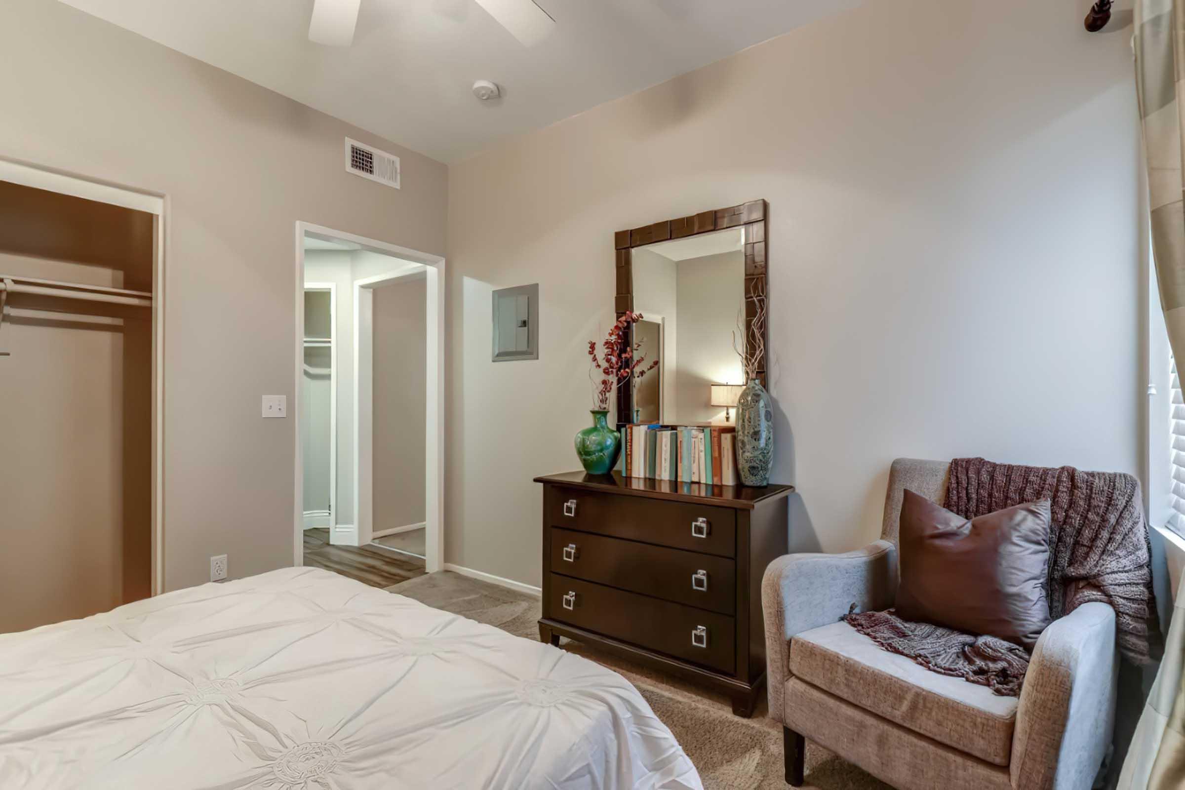 A cozy bedroom featuring a light-colored wall, a neatly made bed with a decorative quilt, a stylish dark wooden dresser with books and a vase, a comfortable armchair with pillows, and a mirror above the dresser. A closet door is partially visible, and there's natural light coming through a window.