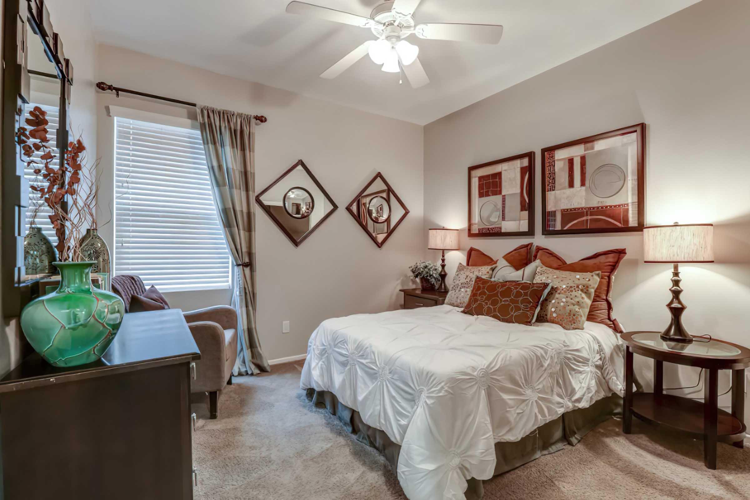 A cozy bedroom featuring a neatly made bed with decorative pillows, a bedside table with a lamp, a dresser, and a comfortable chair. Natural light filters through window blinds, and decorative mirrors are mounted on the wall, adding a touch of elegance to the space. Soft color tones create a warm atmosphere.