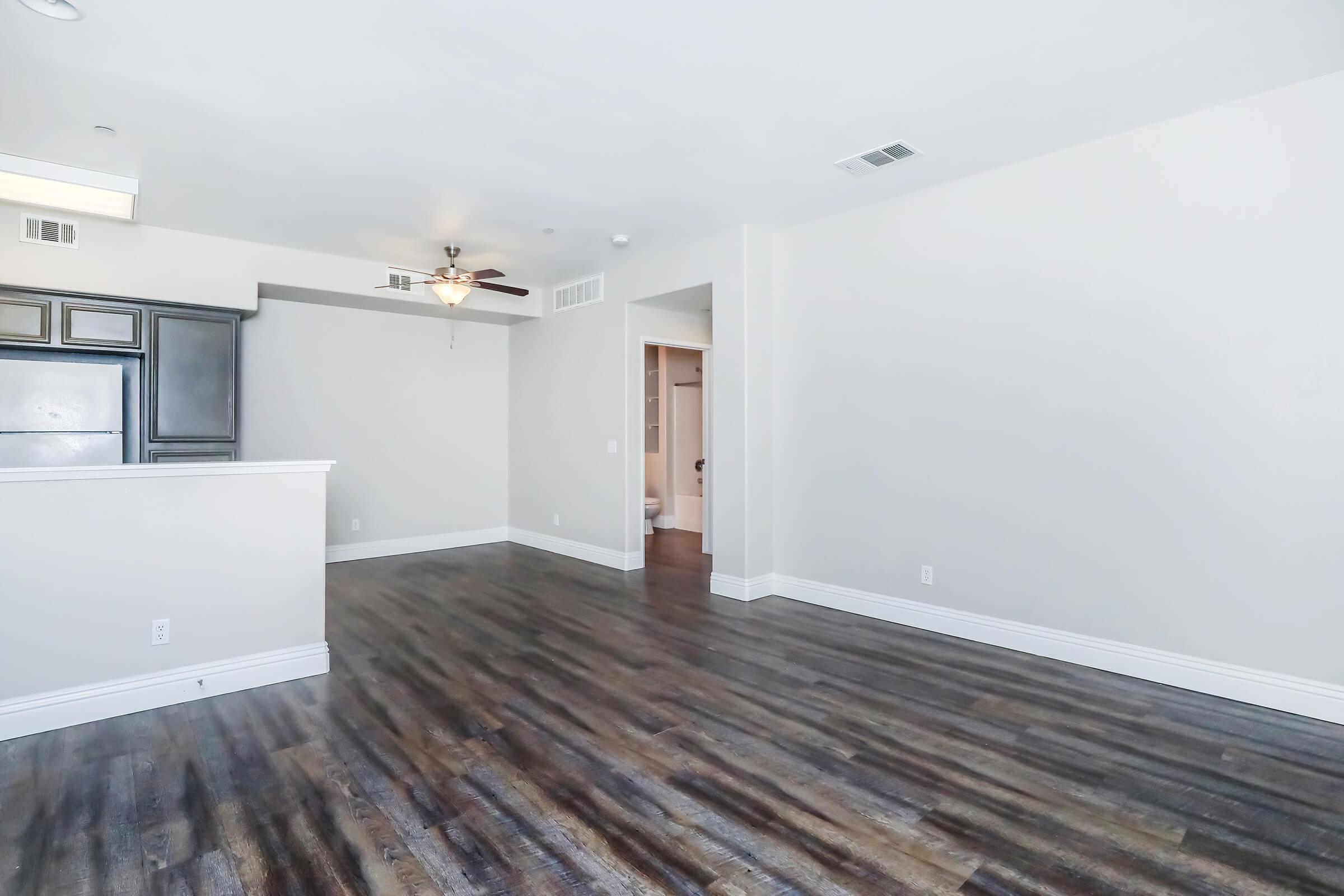 Spacious room with light-colored walls and modern flooring. An open layout features a ceiling fan and a view of a kitchen area with dark cabinetry. A doorway leads to another room at the back, while a bathroom door is visible in the background. Bright and airy atmosphere.