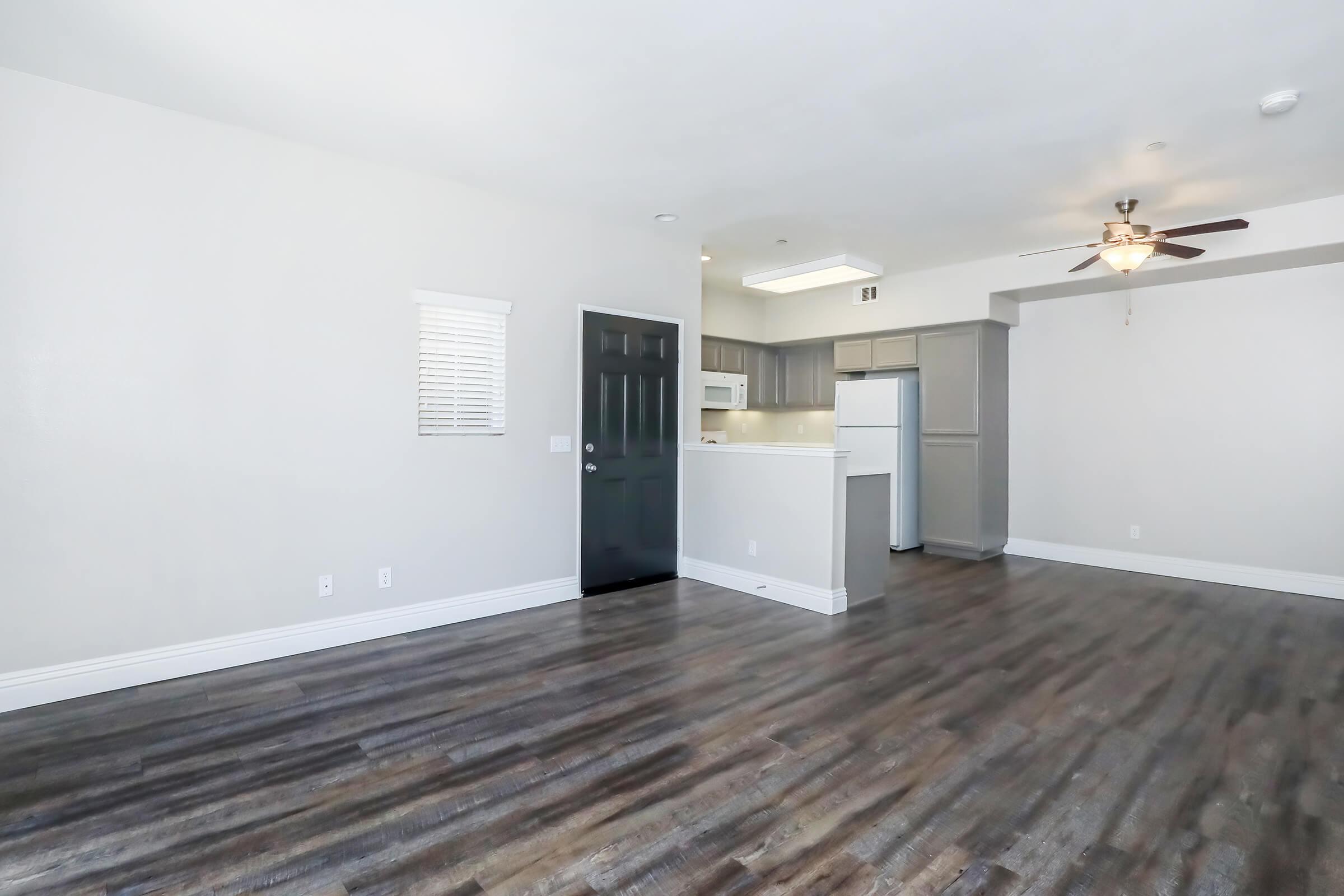 A spacious living area with gray walls and brown laminate flooring, featuring a ceiling fan. On the left, there's a doorway leading to another room. In the background, a kitchen area with white appliances and cabinets is partially visible. Natural light enters through a small window.