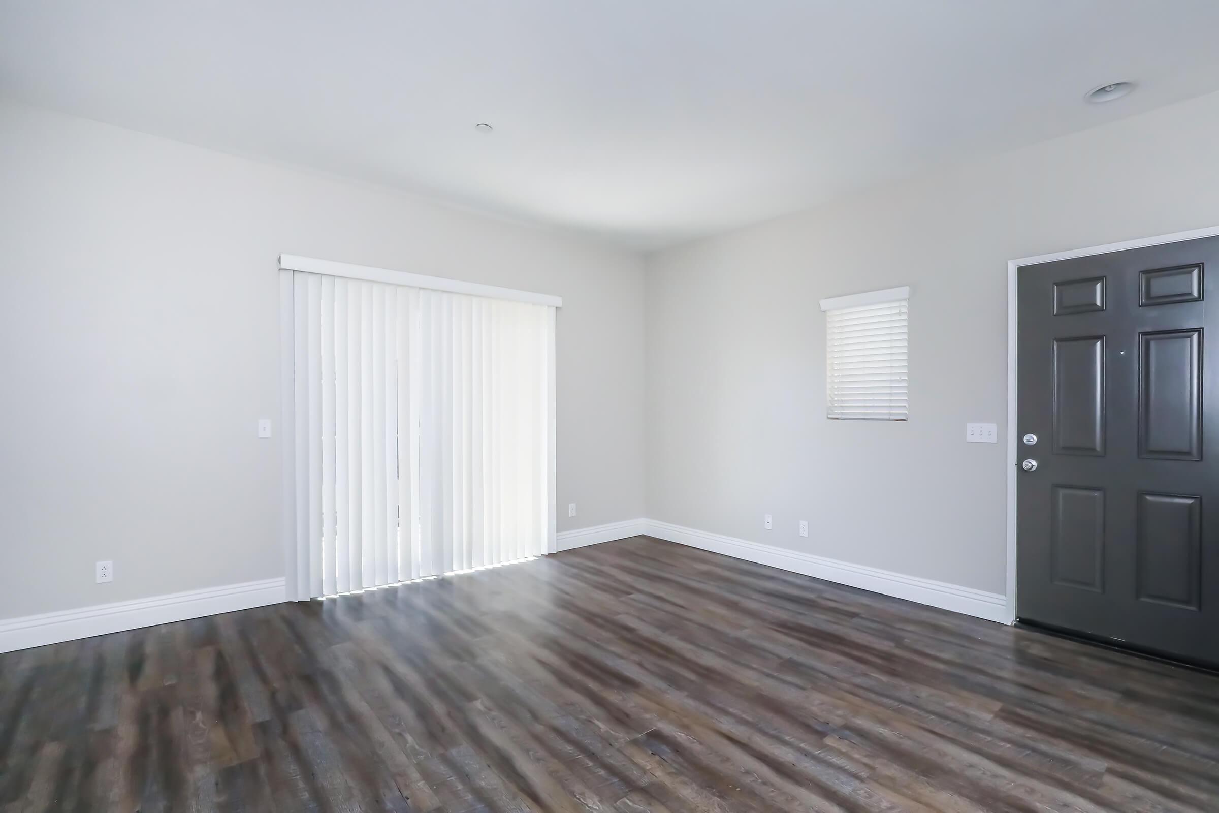 An empty room with light gray walls and wood laminate flooring. A sliding glass door with vertical blinds allows natural light, and a window with blinds is visible on one wall. The entrance features a dark colored door, and there are no furnishings in the space.