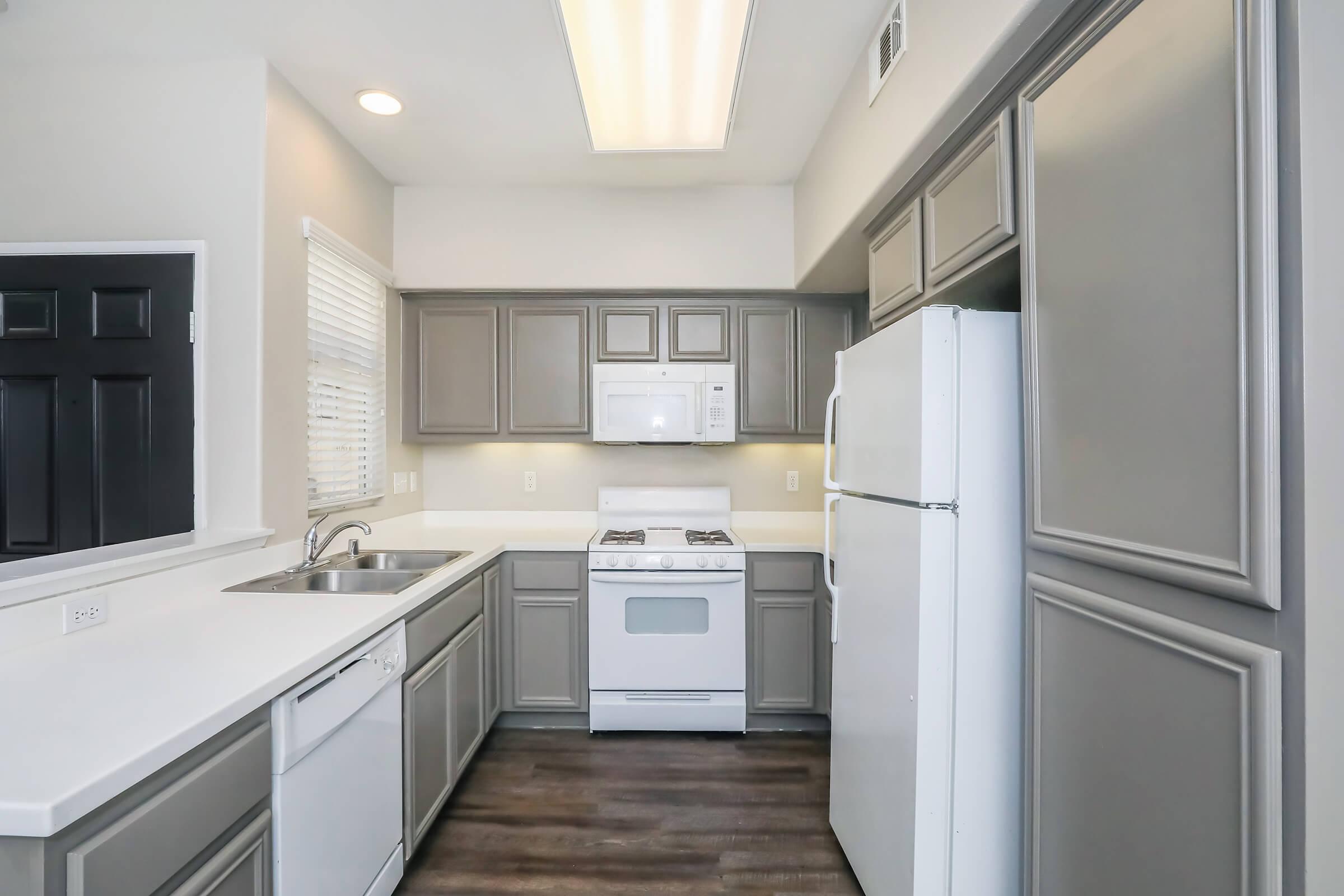 A modern kitchen with gray cabinets, a white countertop, a stainless steel sink, a gas stove, and a built-in microwave. The refrigerator is white, and there is a window with blinds. The flooring is dark wood, creating a contemporary look. Lighting is bright from a ceiling fixture.