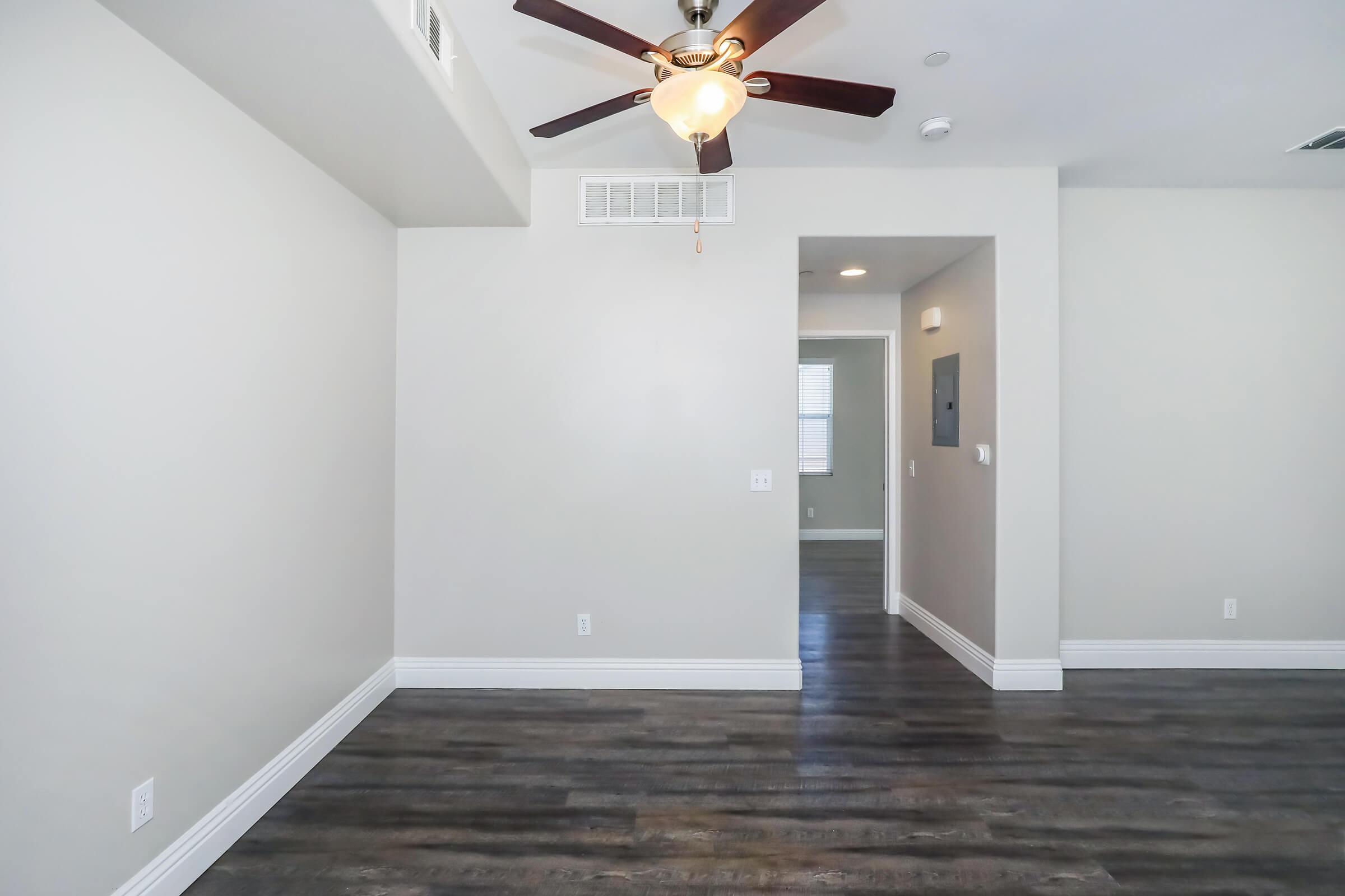 Spacious interior of a room featuring light gray walls, a ceiling fan with a light fixture, and hardwood-style flooring. An archway leads to a hallway, with a small wall panel visible for electrical controls. Natural light enters through a window, enhancing the bright and airy atmosphere.