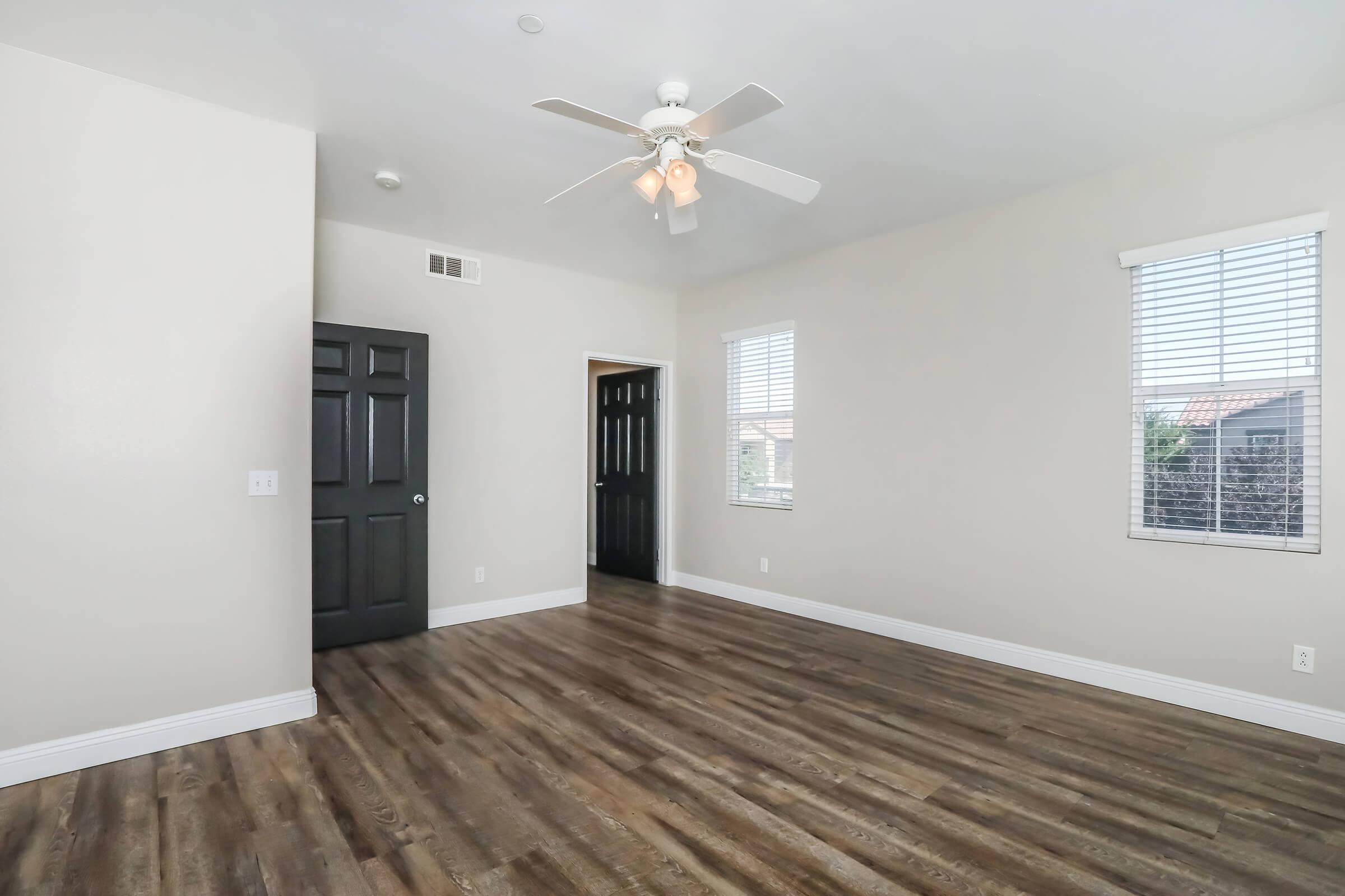 A spacious, well-lit room with light gray walls and dark wood flooring. It features a ceiling fan, two windows with white blinds, and a black double door leading to another room. The room is empty, creating a clean and open feel.