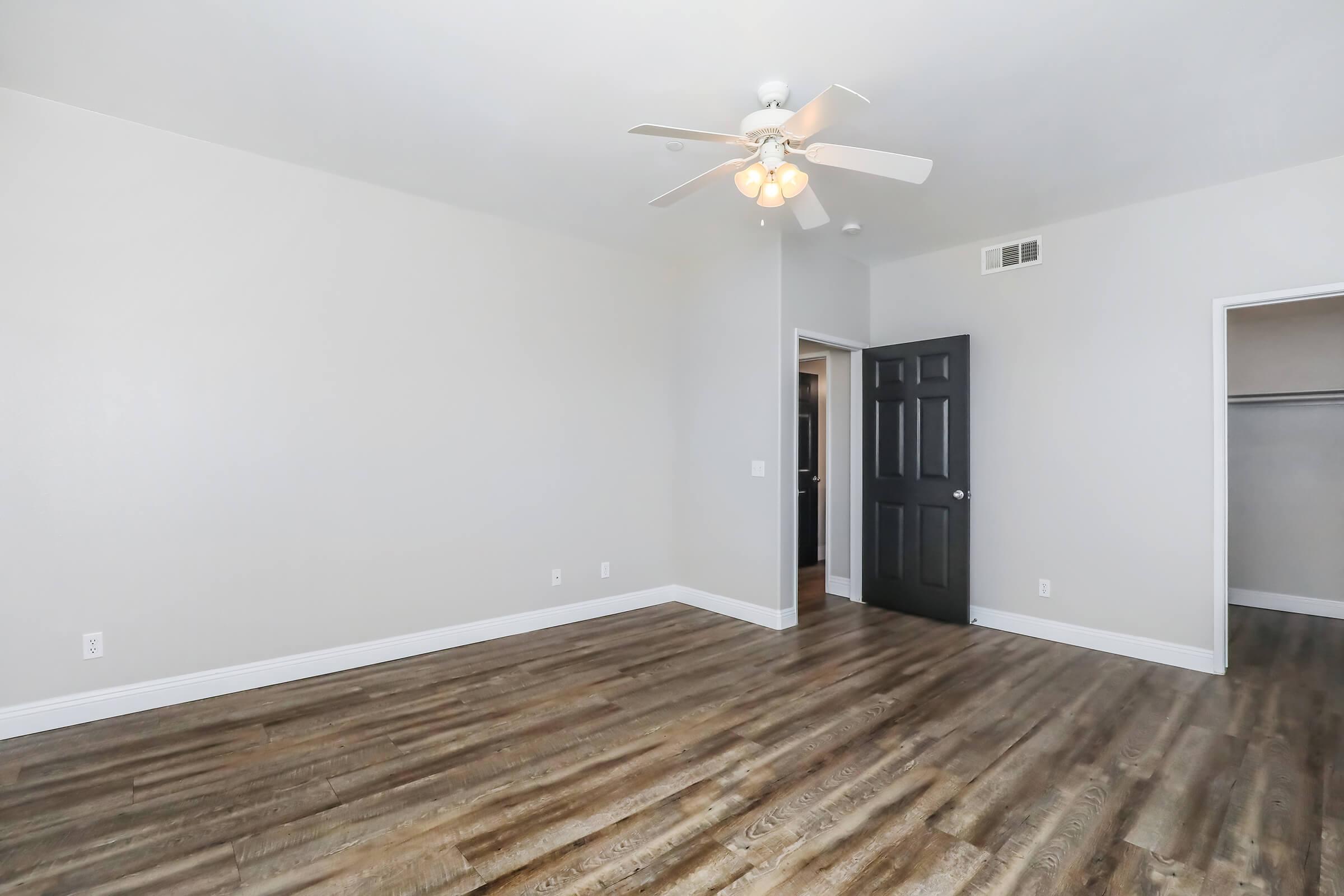 A spacious, well-lit room with a ceiling fan and light fixture. The walls are painted a light color, and there is a door leading to a closet and another room. The floor features hardwood-like planks, contributing to a modern, minimalist aesthetic.