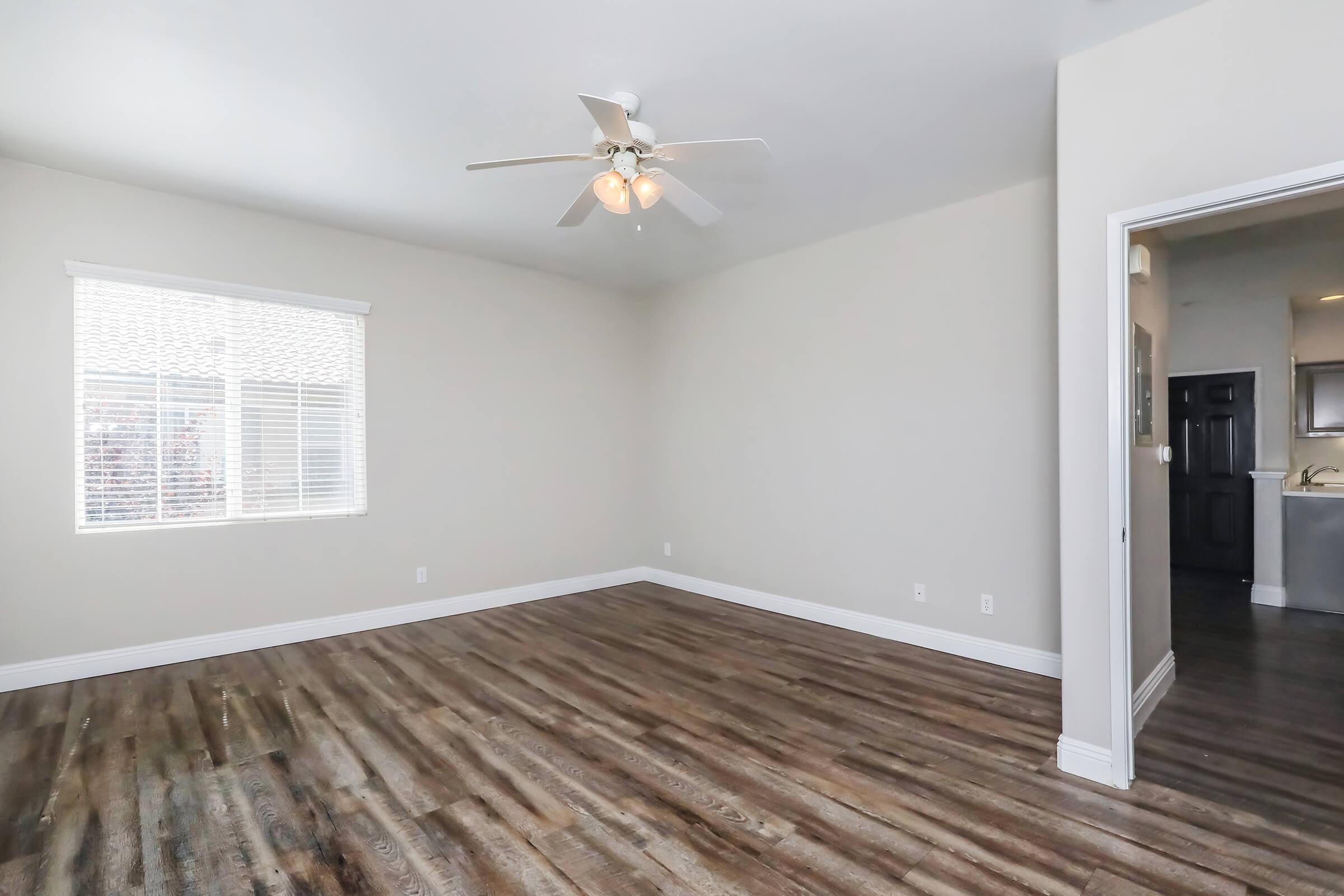 A spacious, empty room with light-colored walls and a ceiling fan. The floor features hardwood-like planks. A window on the left allows natural light, and a doorway on the right leads to another area, possibly a kitchen.