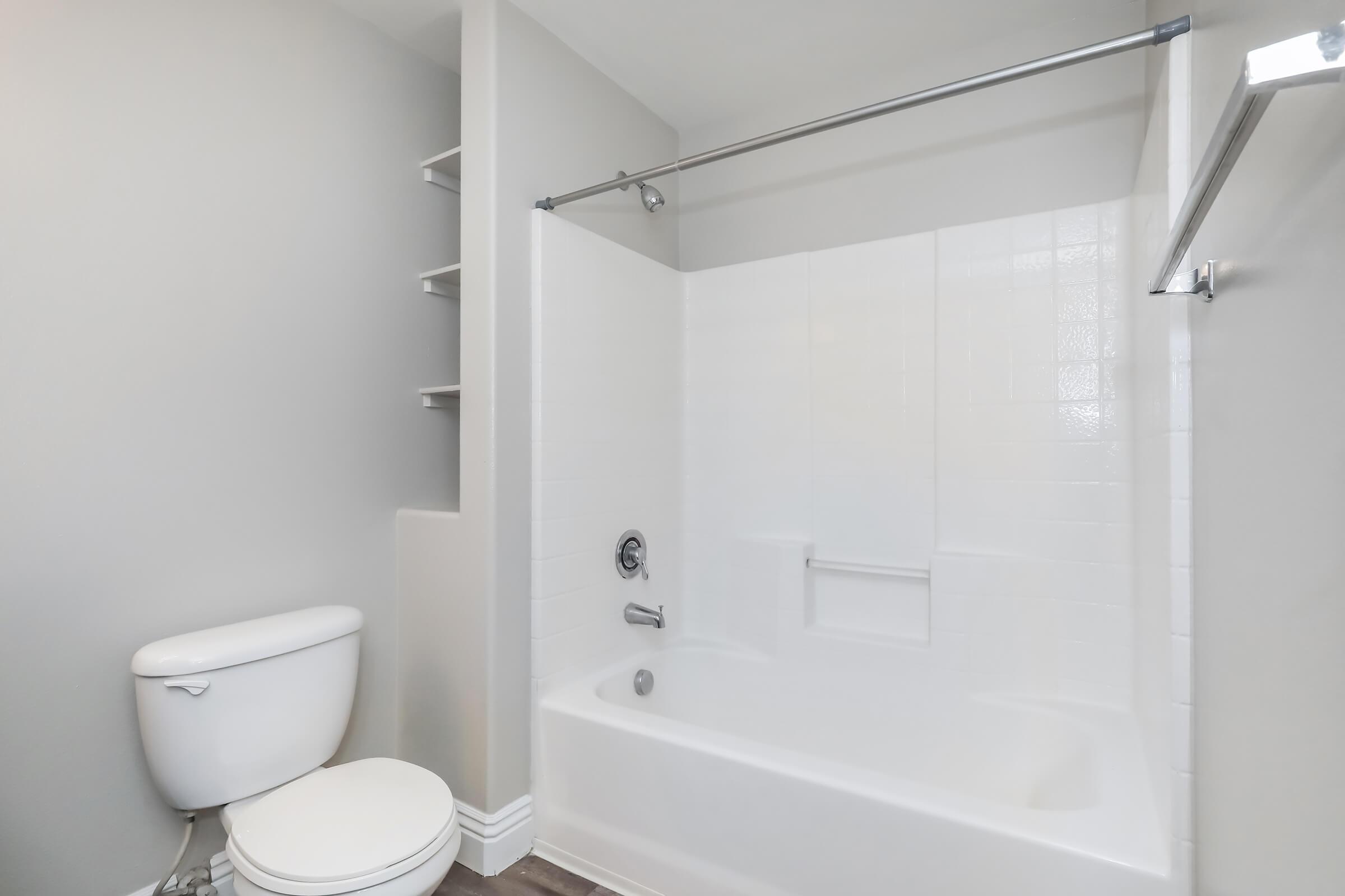 A clean and modern bathroom featuring a white bathtub and shower combination, a white toilet, light gray walls, and a small shelf on the side. The space is well-lit with a minimalist design.