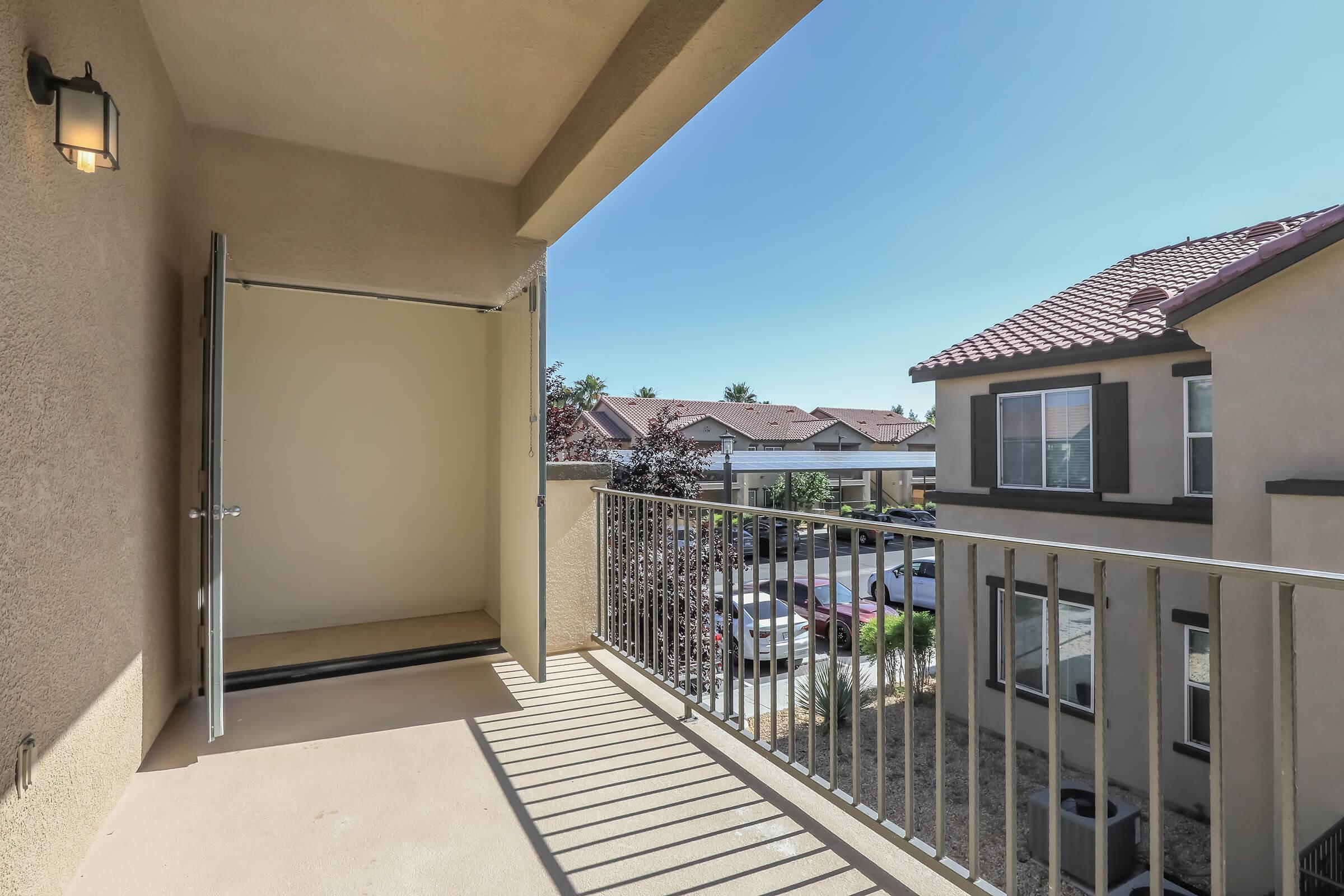 A view of a balcony with an open door, featuring a railing and a clear sky. The scene includes adjacent buildings with red-tiled roofs and parked cars below, creating a sunny, residential atmosphere. Shadows cast on the balcony floor add depth to the image.