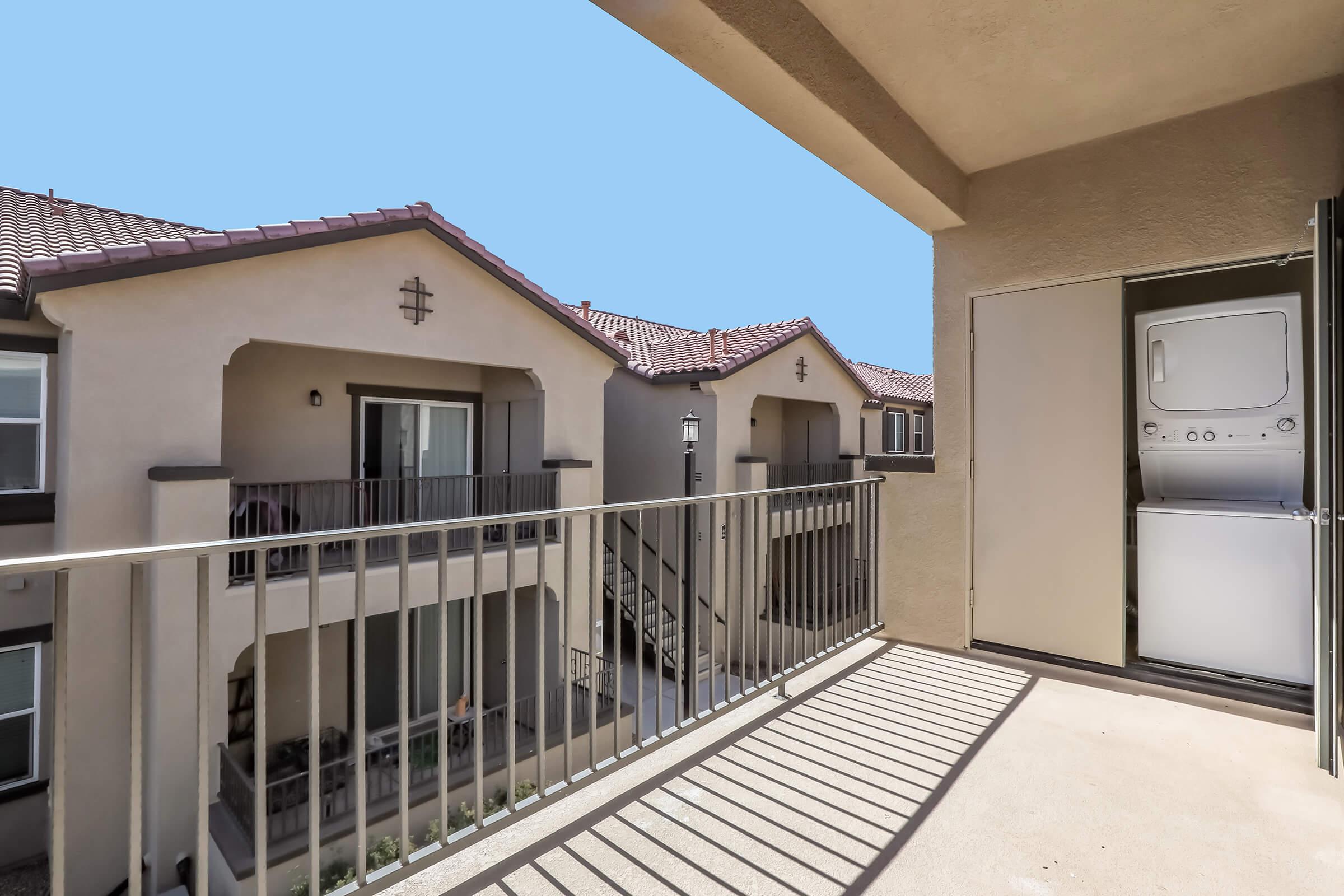 A balcony view of an apartment complex featuring two buildings with Mediterranean-style architecture. The balcony includes a railing, with a stackable washer and dryer positioned nearby. The sky is clear and blue, enhancing the cozy outdoor space.