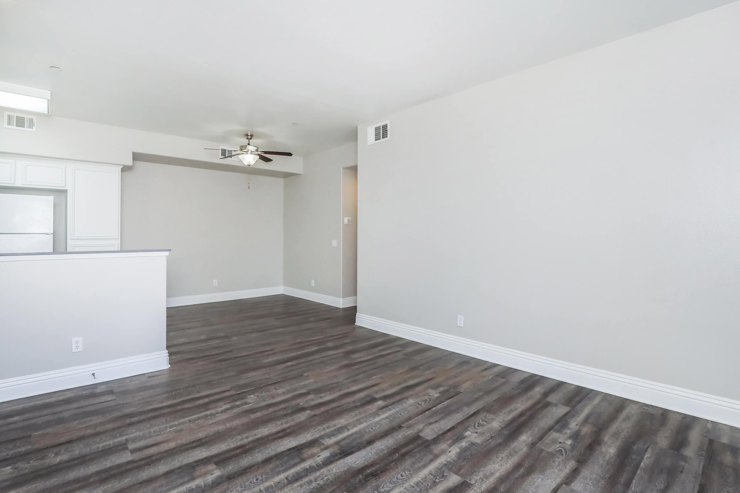 Empty living area with light gray walls and a wooden floor. A ceiling fan is visible in the center of the room. There is a partial wall, possibly leading to a kitchen area, on the left. A doorway is seen on the right, leading to another room or hallway. The space has plenty of natural light.