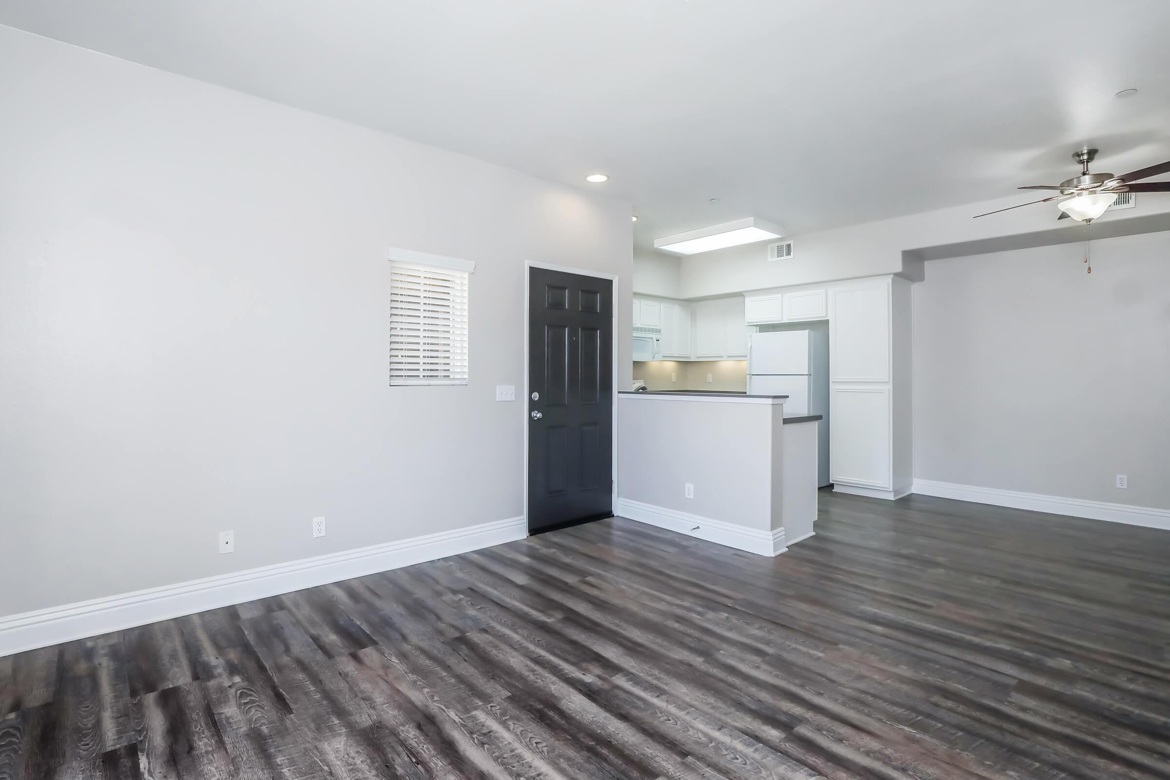 Spacious living area with grey walls, dark wood flooring, and a ceiling fan. A black door leads to the entrance, and a kitchen is visible in the background with light-colored cabinetry. Natural light filters in through a small window. The layout contributes to an open, inviting atmosphere.