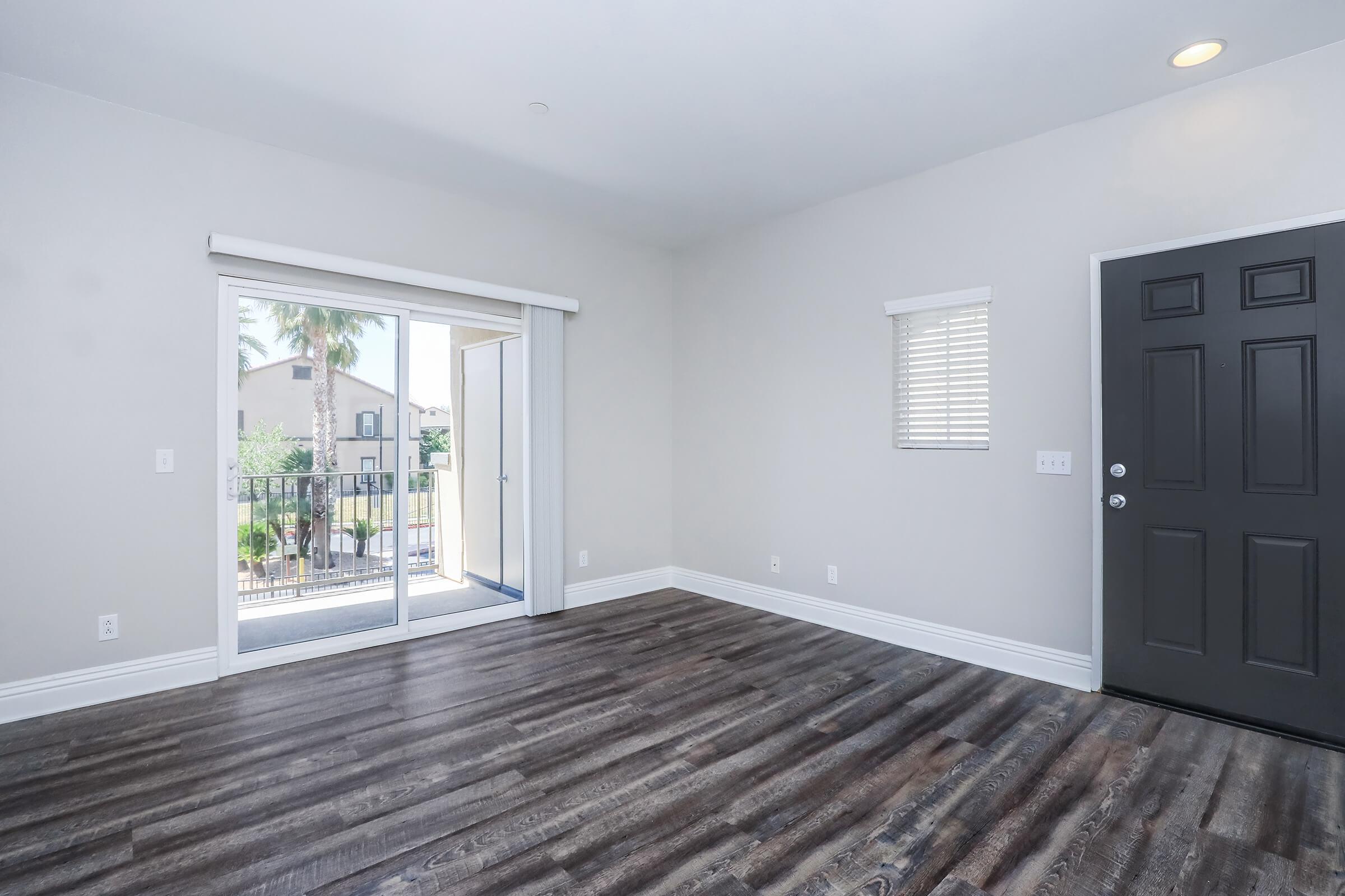 Spacious, empty room featuring light-colored walls, modern flooring, and a sliding glass door that opens to a balcony. A dark front door is visible, along with a small window and natural light coming in, creating a bright and inviting atmosphere.