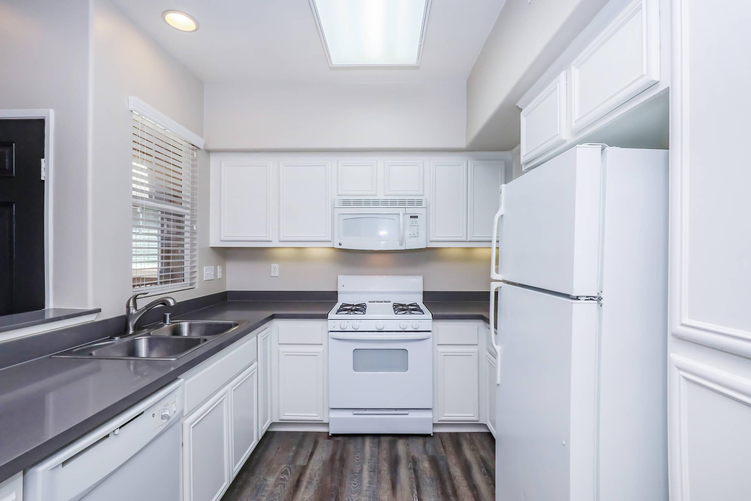 A modern kitchen featuring white cabinets, a gray countertop, a double sink, and a white stove and microwave. There is also a white refrigerator, and natural light coming from a window with blinds. The flooring is a dark wood finish.