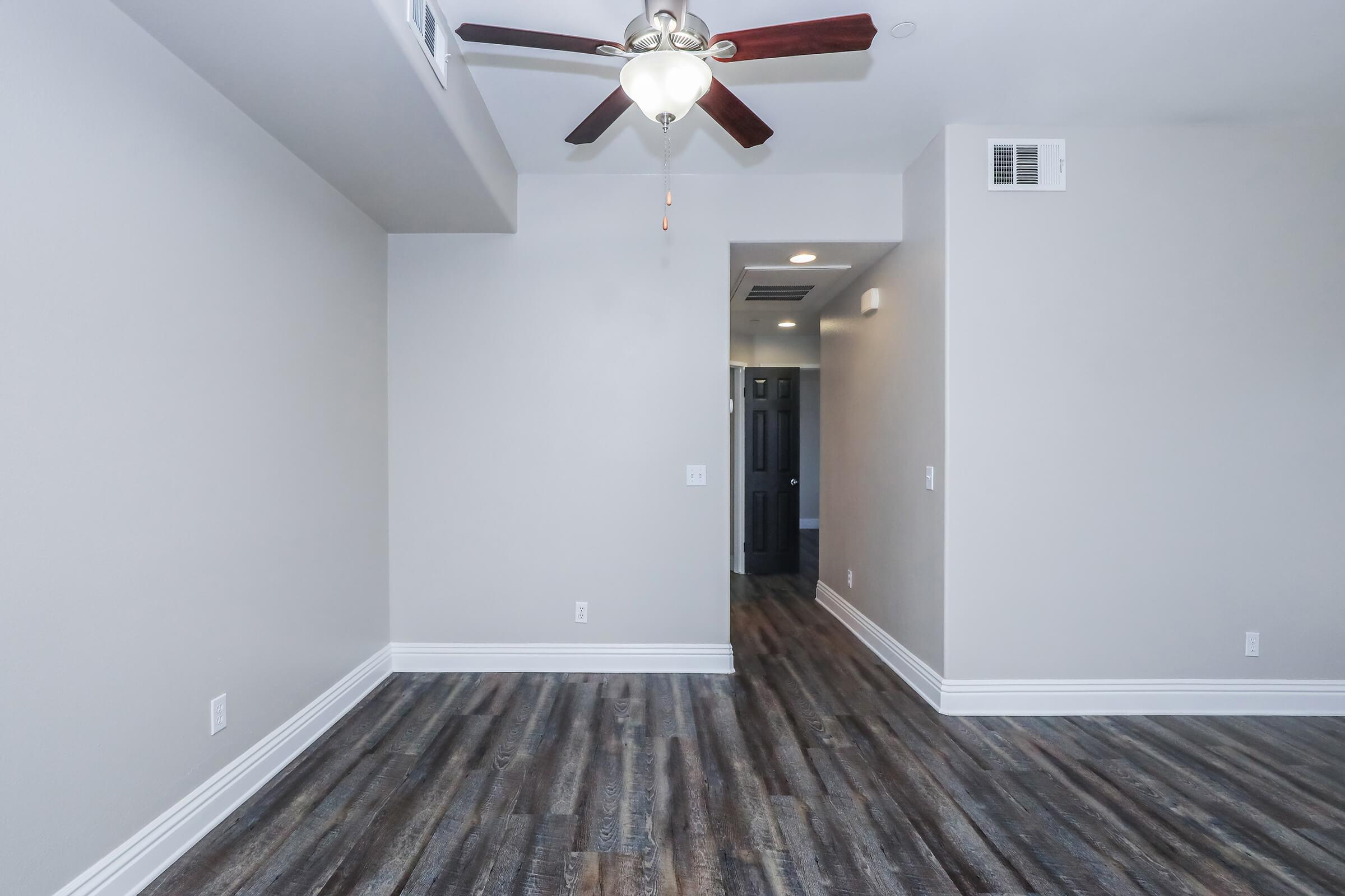 A spacious, empty room with light gray walls and dark wood-like flooring. A ceiling fan with wooden blades is centered overhead. To the right, a doorway leads to another area. Natural light enters through a small opening, highlighting the clean and modern design.