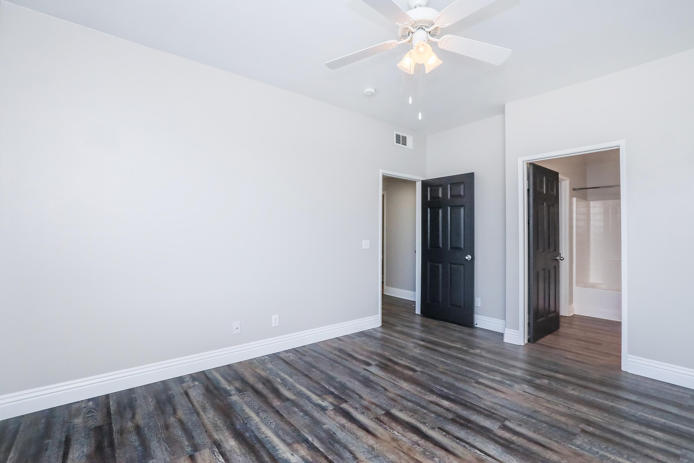 Empty room with light gray walls and dark wooden flooring. A ceiling fan is mounted in the center. Two doors are visible: one leading to an adjoining space and another possibly to a bathroom. Natural light fills the room, enhancing the spacious feel.